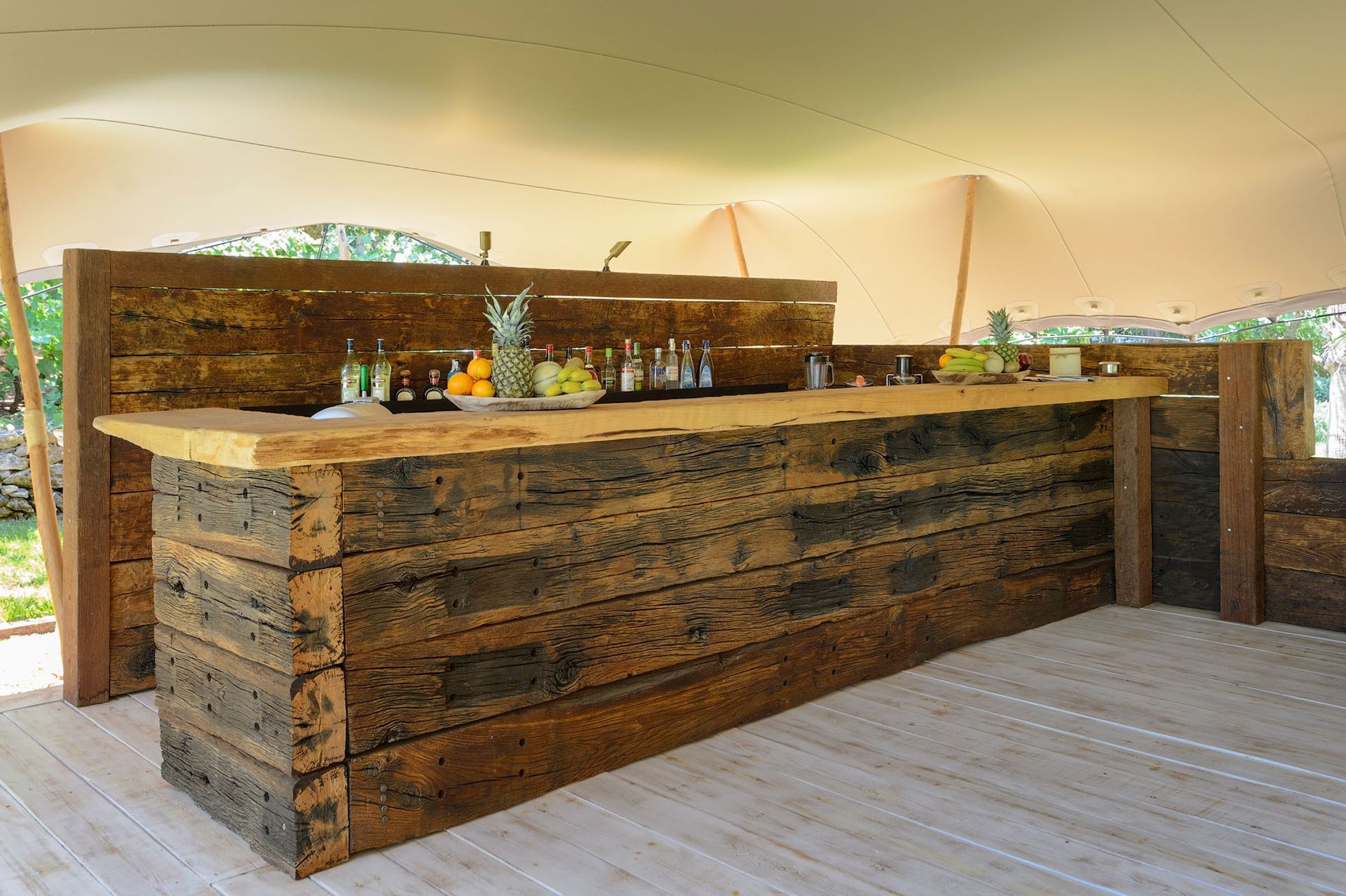 Barra de madera con bebidas y frutas bajo una carpa.