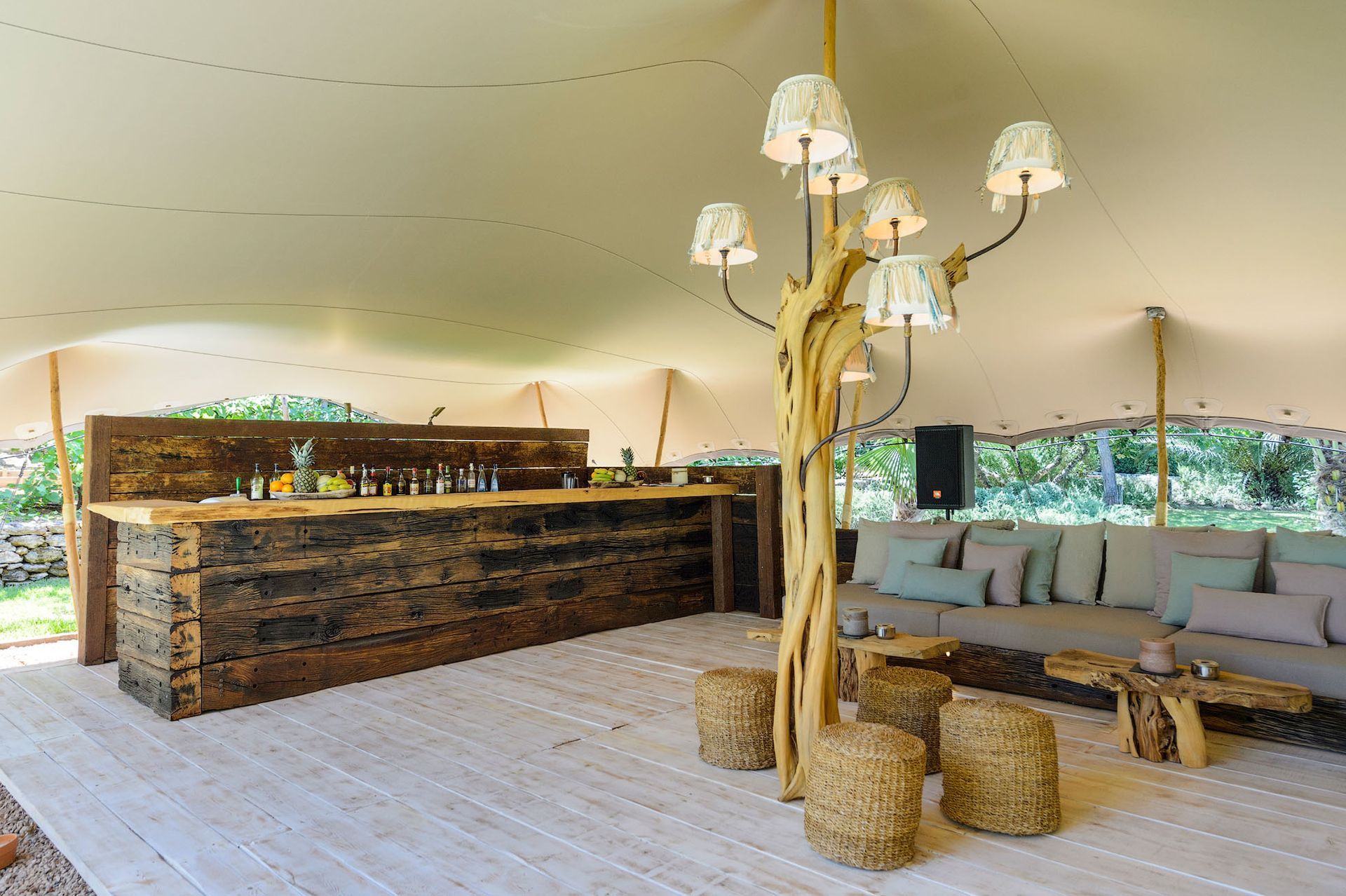 Zona de estar al aire libre bajo una carpa, con bar y asientos. Barra rústica de madera, lámpara con forma de árbol