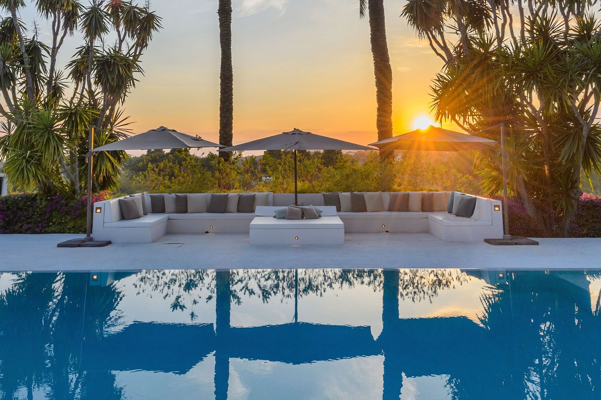 Salón junto a la piscina con sombrillas y la puesta de sol reflejándose en el agua.