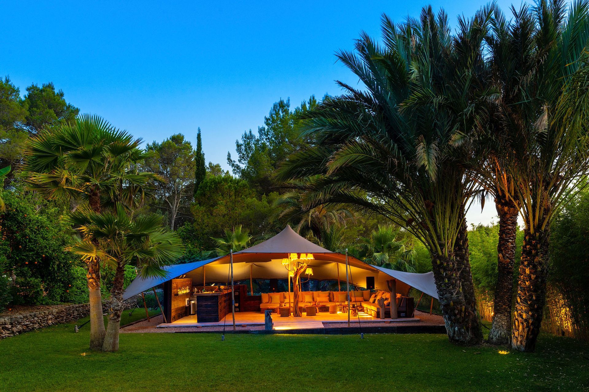 Bar al aire libre con carpa, detalles de madera y un techo alto en punta, ubicado entre exuberantes árboles verdes y césped.
