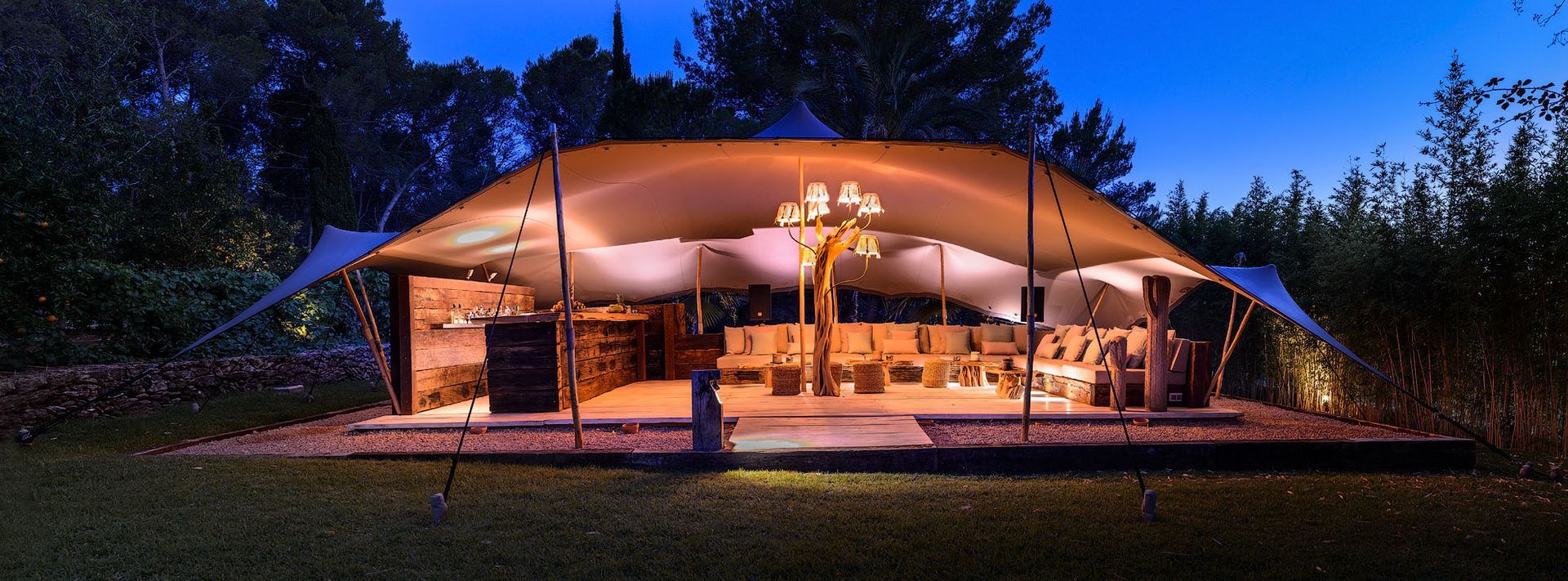 Una carpa instalada para un evento, iluminada por la noche, con mesas y sillas en el interior.