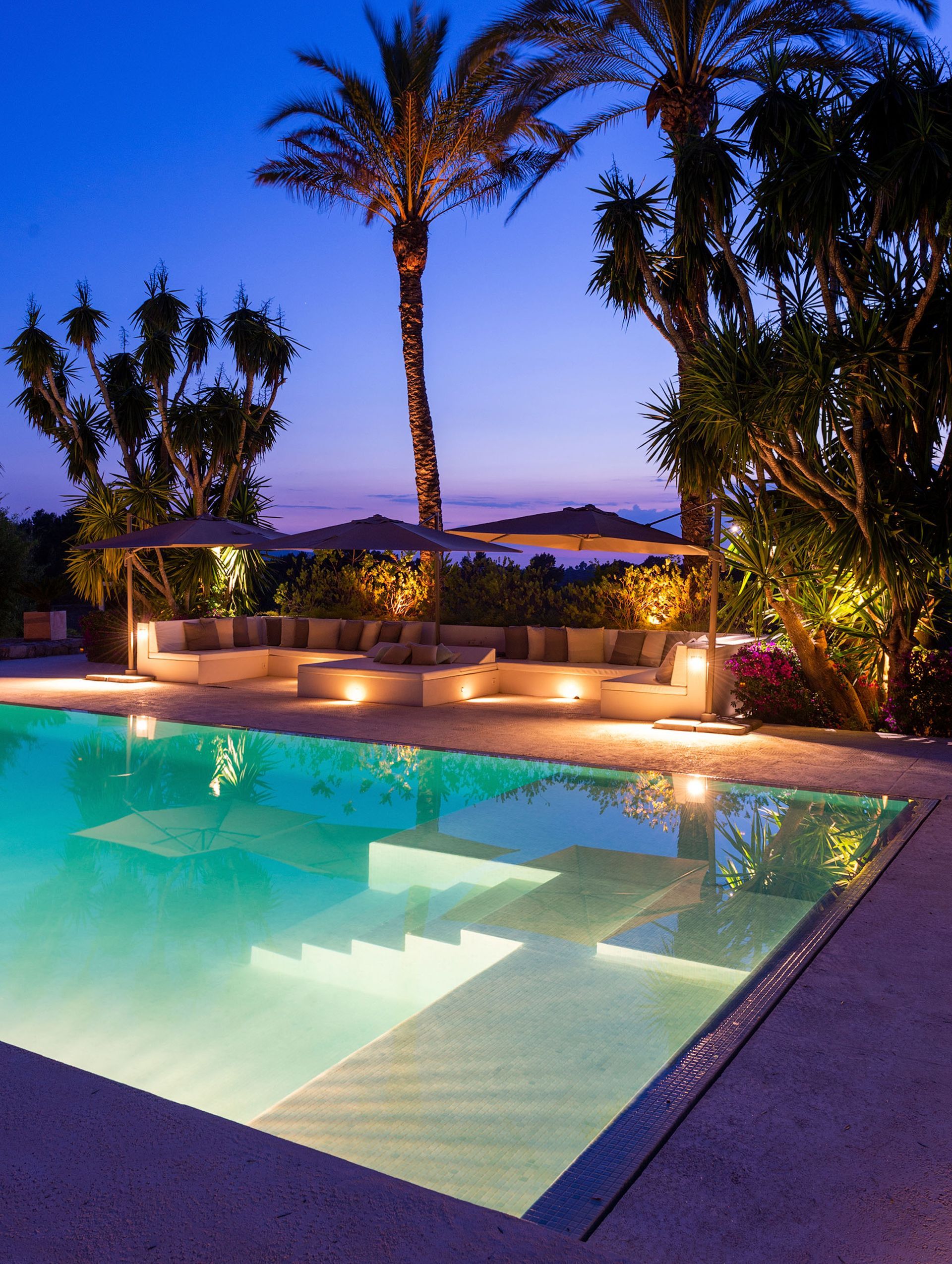 Una vista nocturna de una piscina con escalones incorporados, asientos tipo lounge y palmeras.