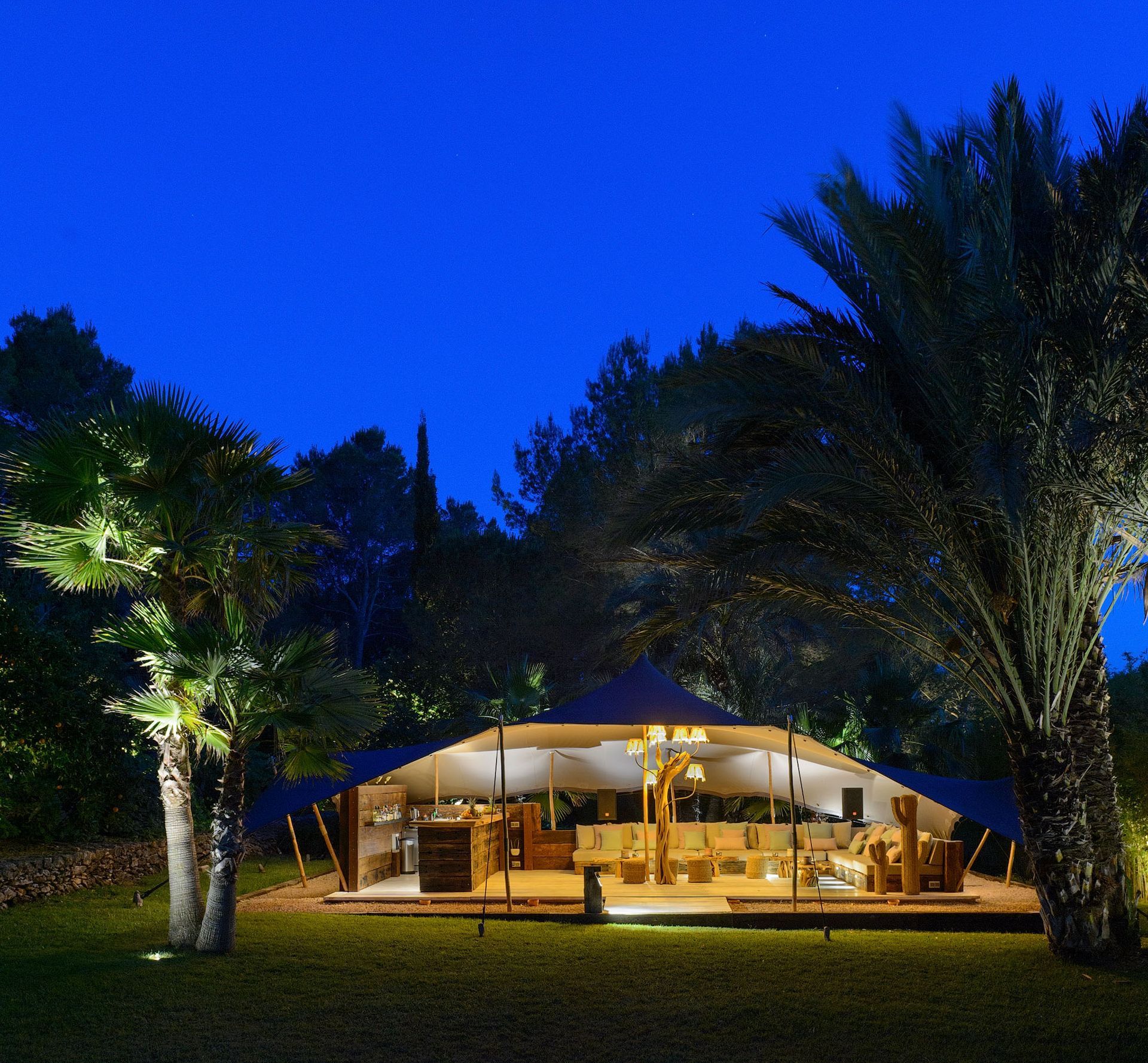 Carpa iluminada al aire libre por la noche, sobre un césped con palmeras. Se ven los muebles del interior. Cielo azul oscuro.