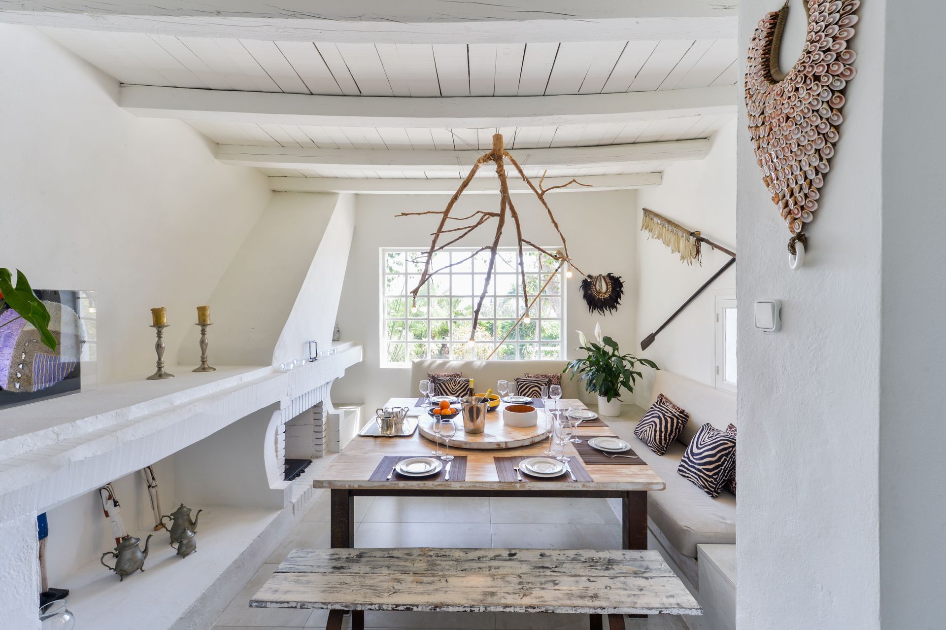 Comedor rústico con mesa de madera, banco y ventana. Paredes blancas y lámpara de araña decorativa con ramas.