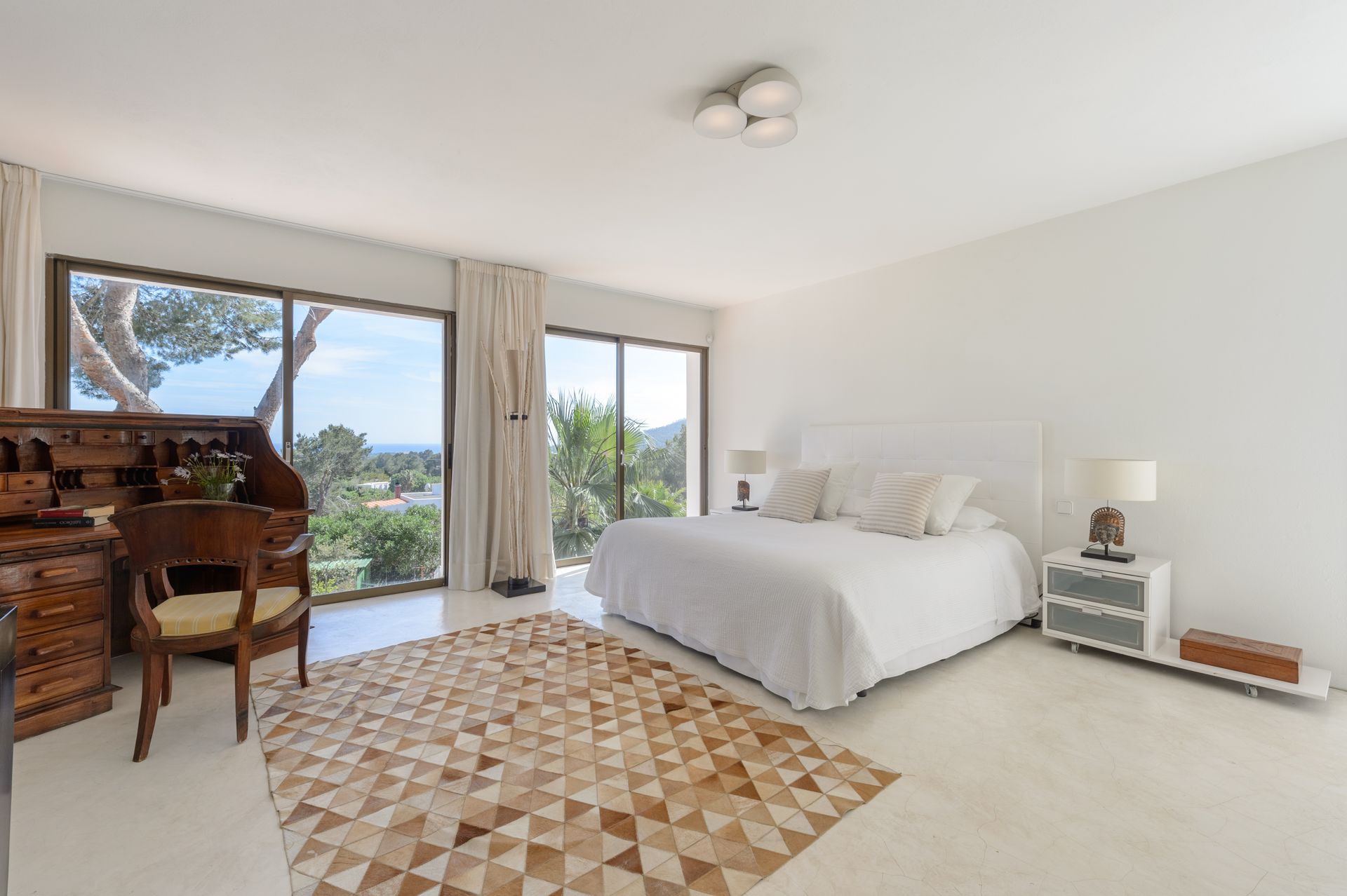Dormitorio con grandes ventanales, cama blanca, escritorio de madera y alfombra con motivos geométricos.