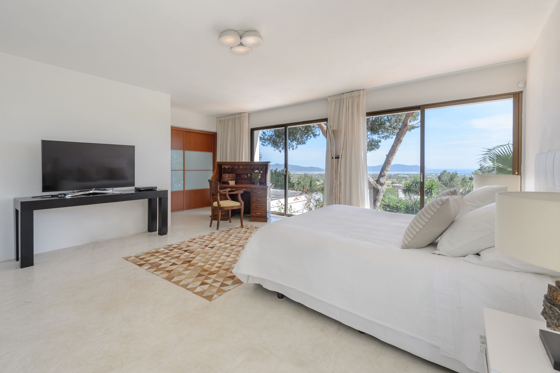 Dormitorio luminoso con cama, TV, escritorio y puertas corredizas de vidrio con vista a los árboles y al mar.