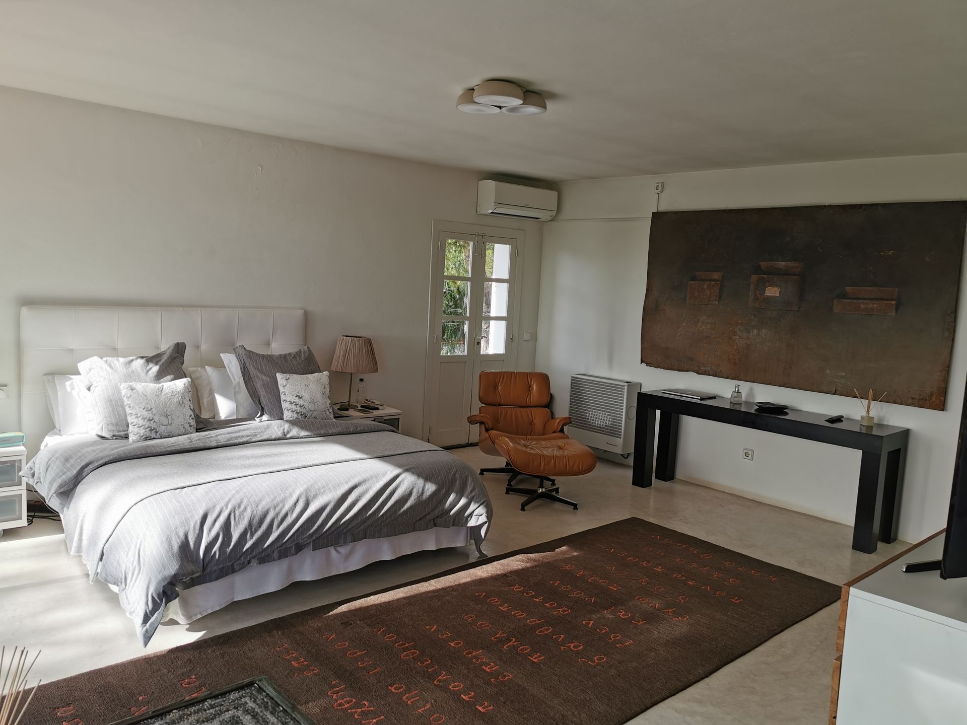 Dormitorio con cama, obras de arte, una silla y una alfombra. La luz del sol entra a raudales por una puerta.