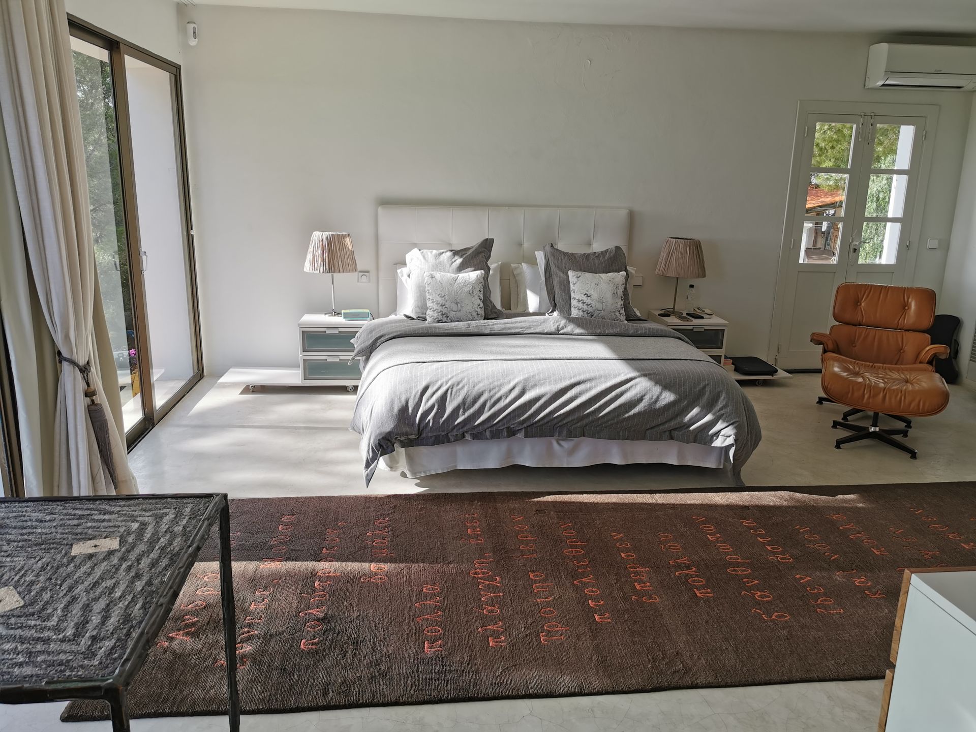 Dormitorio con cama, mesitas de noche, silla y alfombra. La luz natural entra a raudales por un gran ventanal.