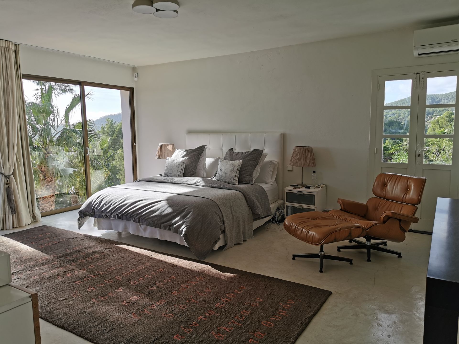 Amplio dormitorio blanco con cama, alfombra marrón, sillón de cuero y gran ventanal con vista a los árboles.