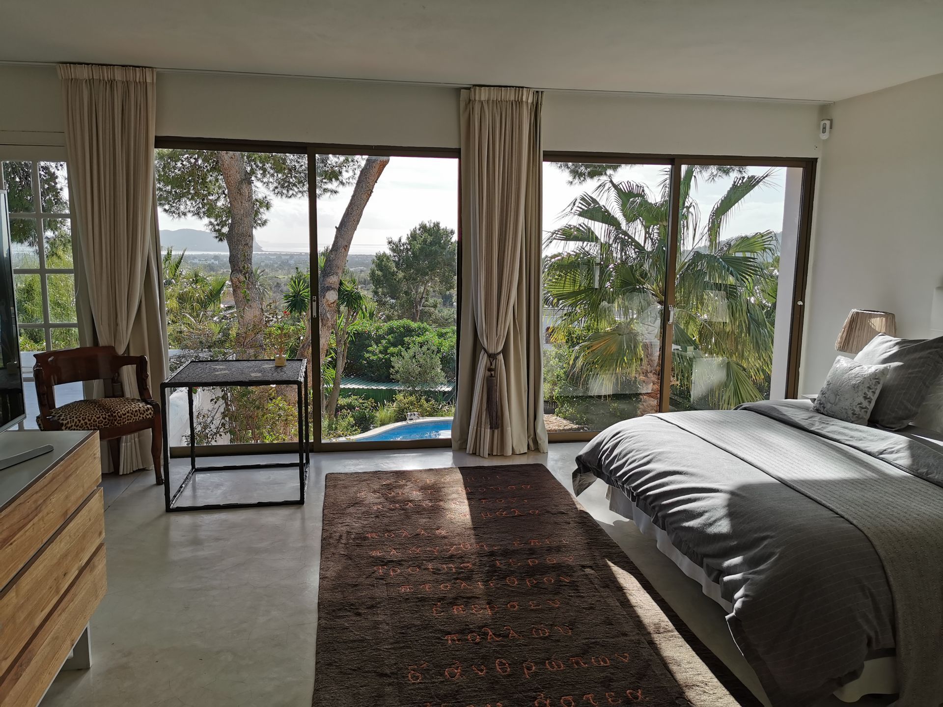 Dormitorio con grandes ventanales, cortinas neutras, una cama y vista a los árboles y a la piscina.