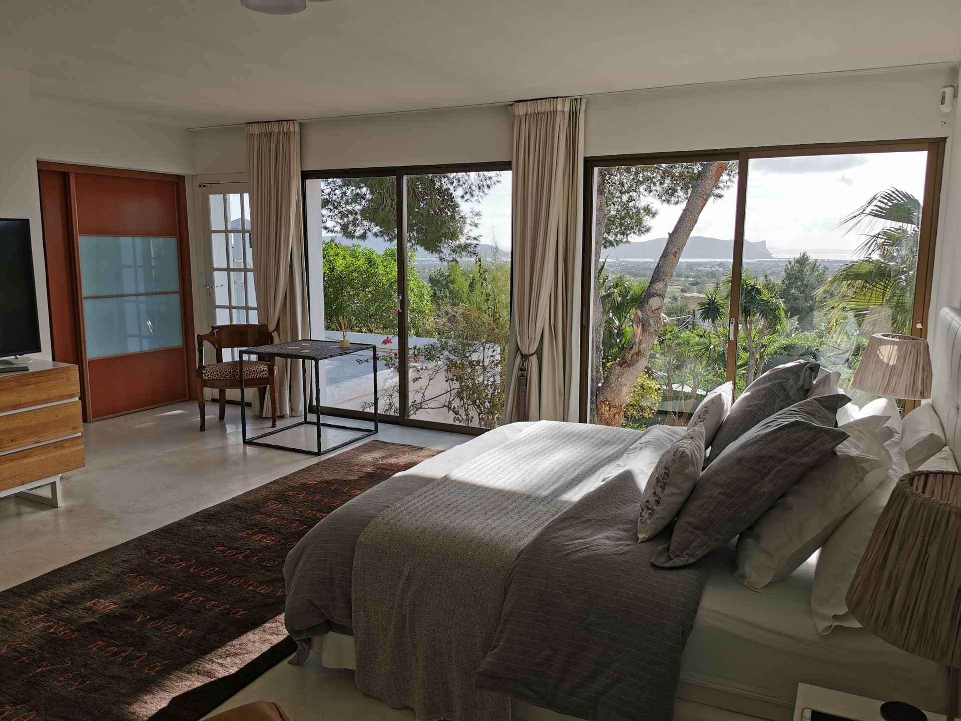 Dormitorio con cama grande, alfombra marrón y ventanas de piso a techo con vista a la vegetación y las montañas.