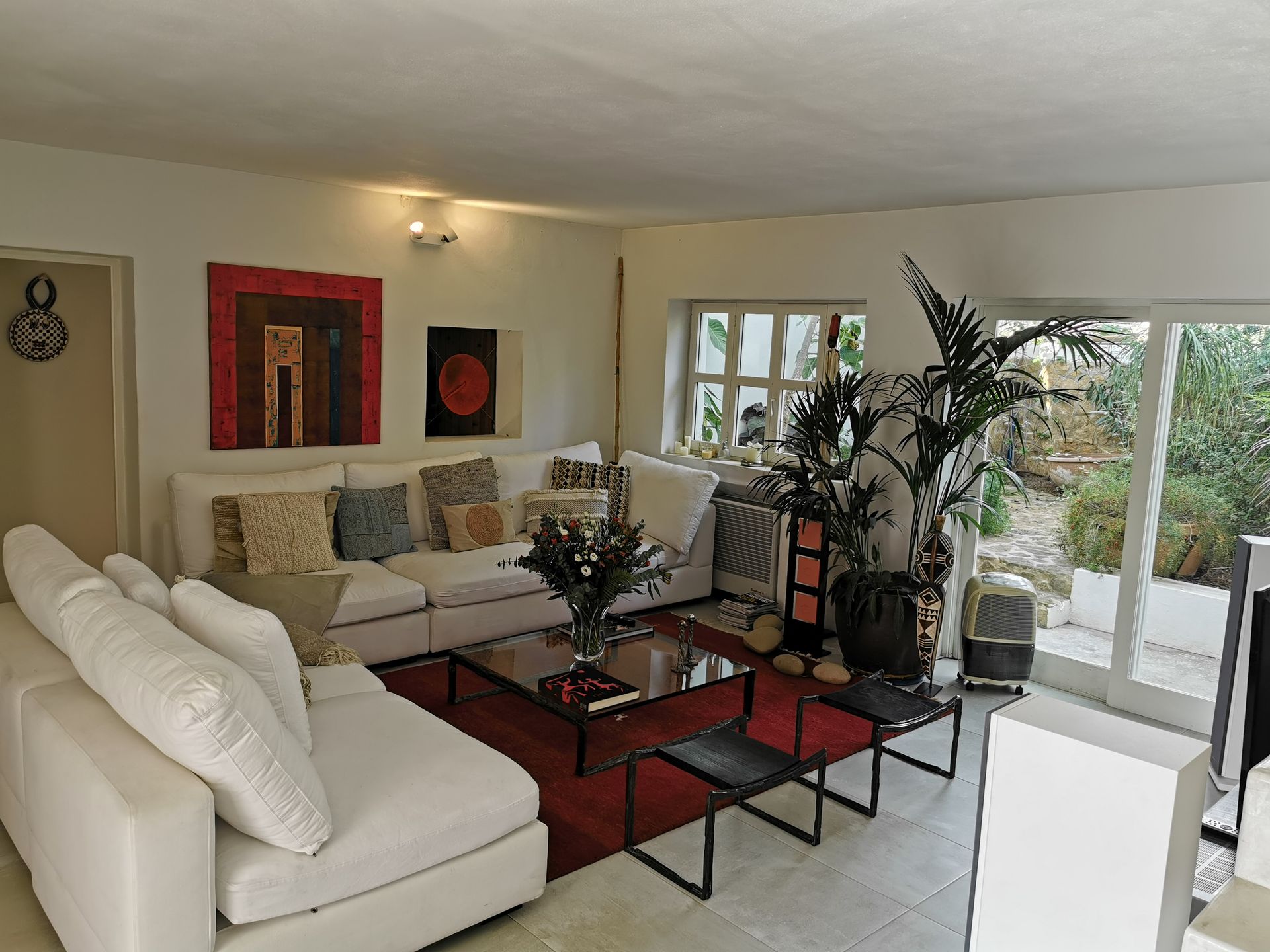 Sala de estar con sofás blancos, alfombra roja, arte, plantas y puertas de vidrio a un jardín.