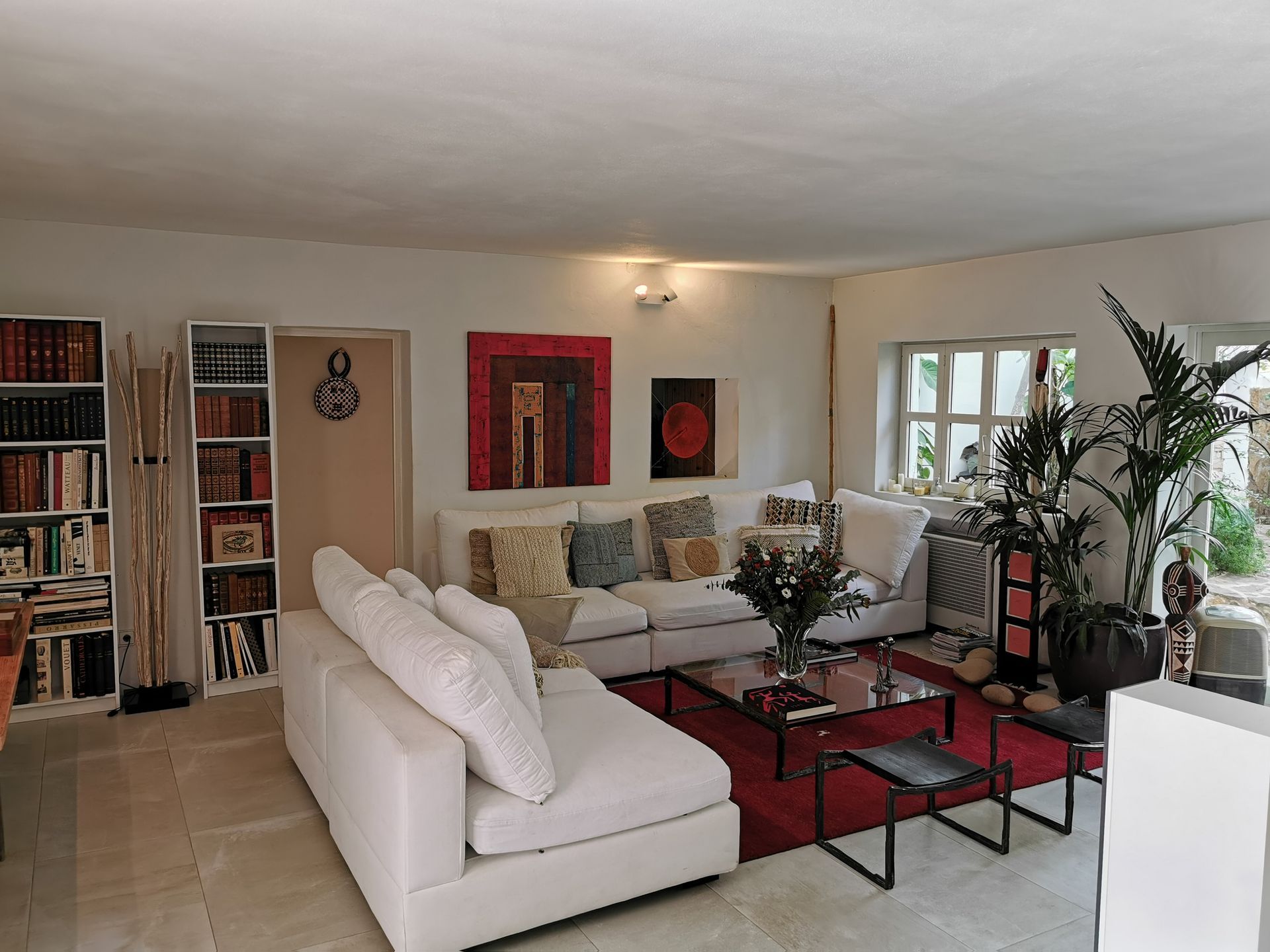 Sala de estar con sofás blancos, estanterías y alfombra roja. Las obras de arte y las plantas le dan color.