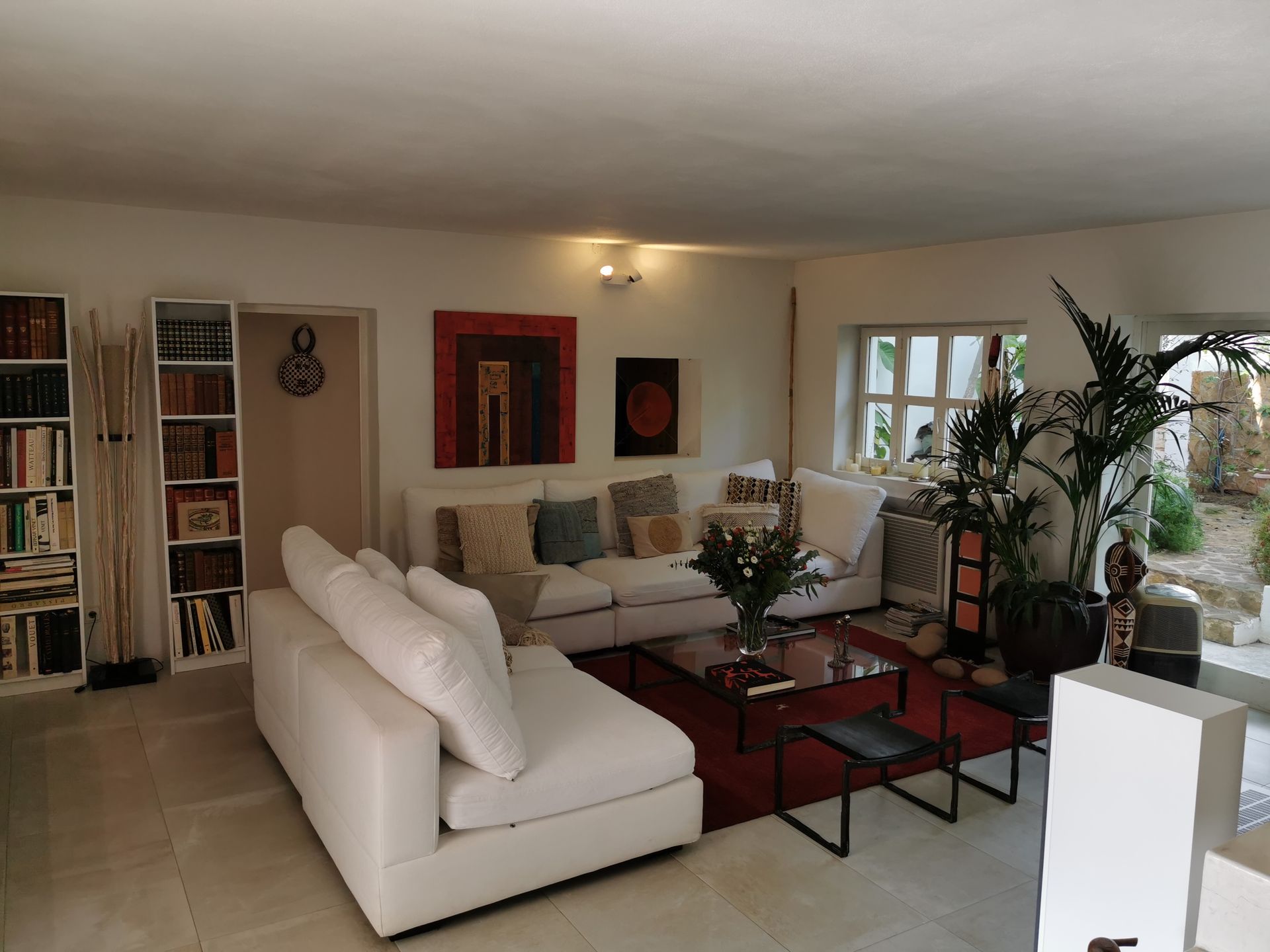 Sala de estar con sofás blancos, estanterías y obras de arte; una alfombra roja y una planta grande.