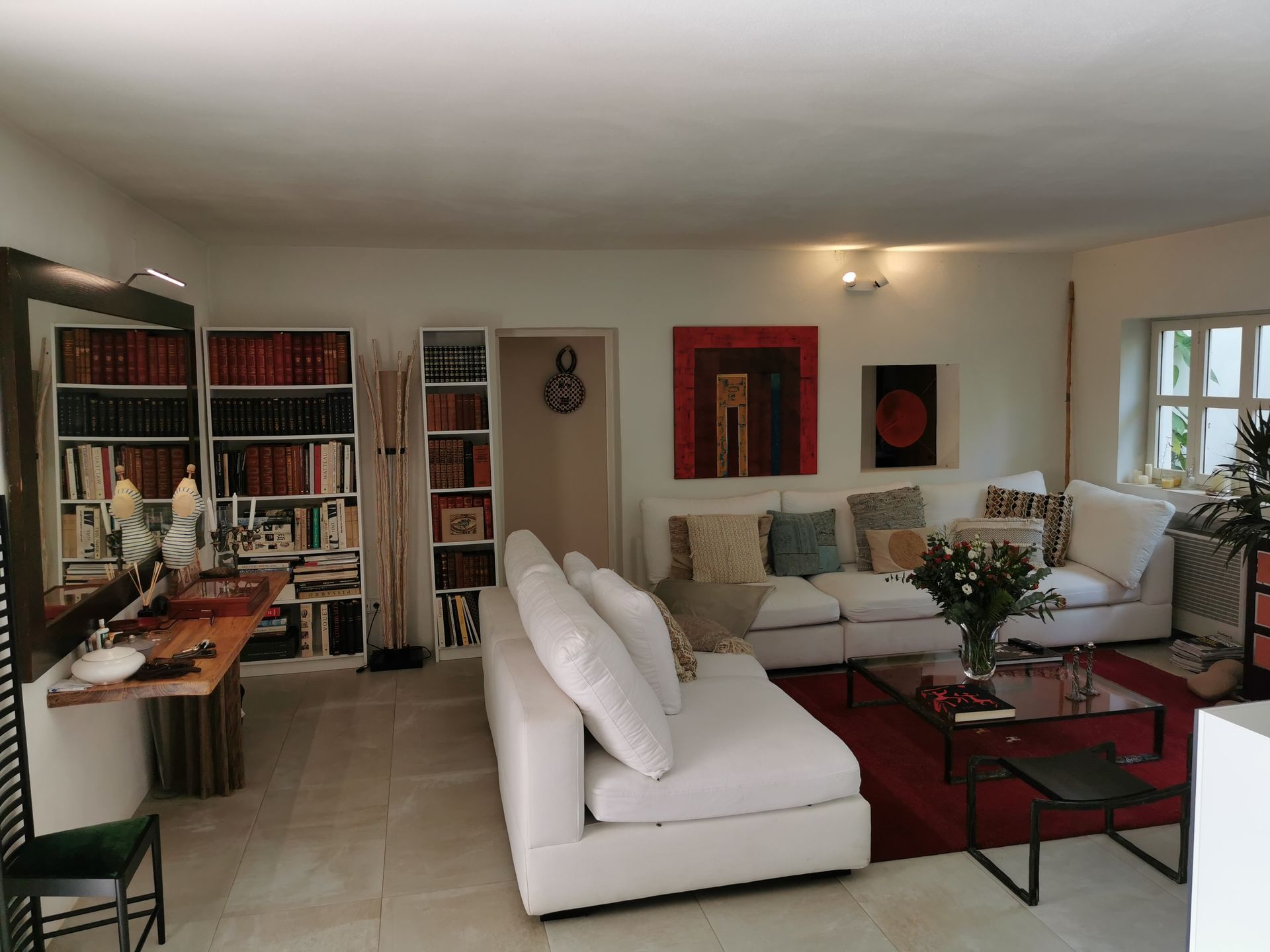 Sala de estar con sofás blancos, estanterías y obras de arte; una alfombra roja y una mesa de café de cristal