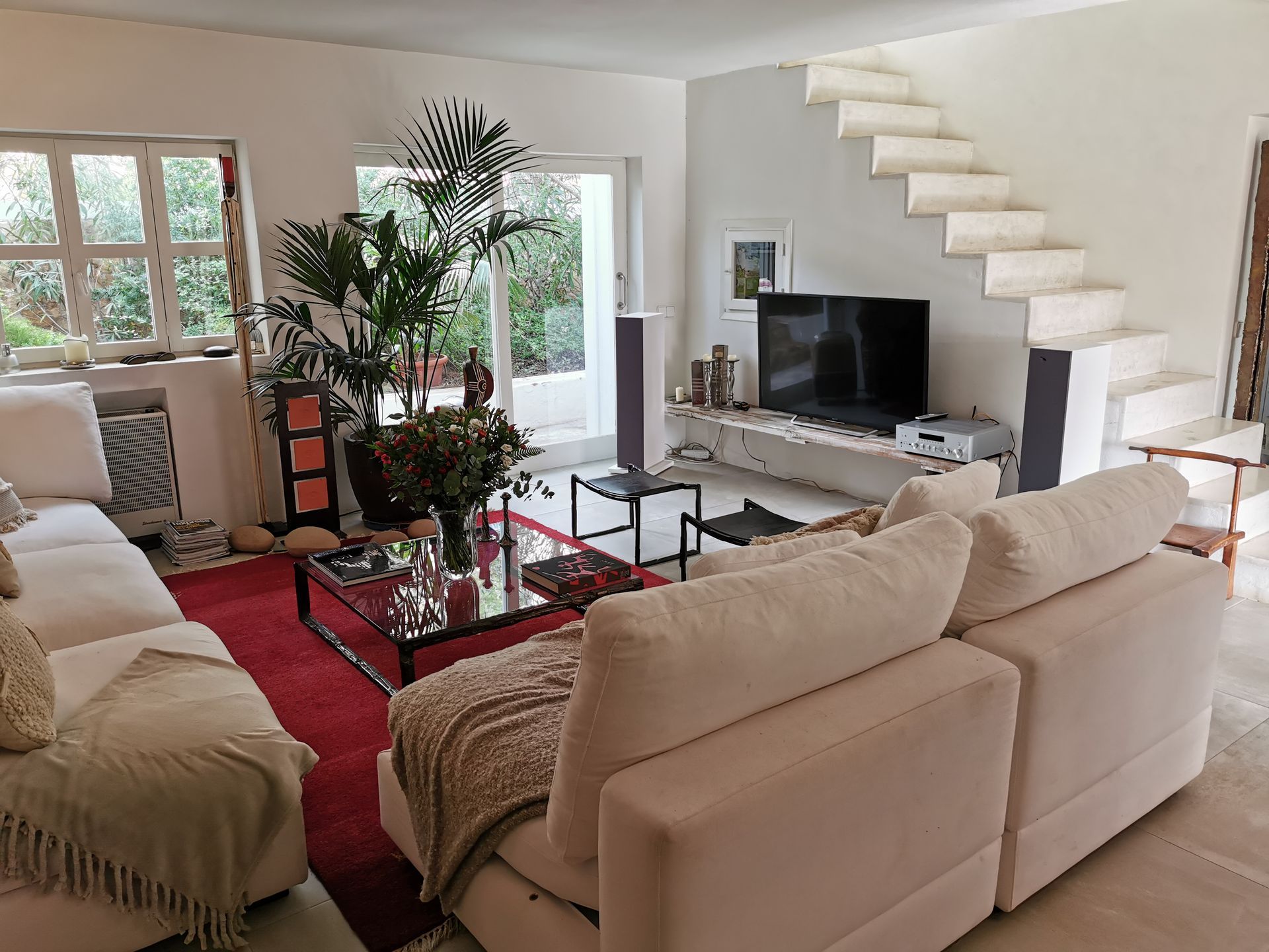 Sala de estar con sofás blancos, alfombra roja, mesa de centro de cristal, televisor y escalera. Gran ventanal y plantas.