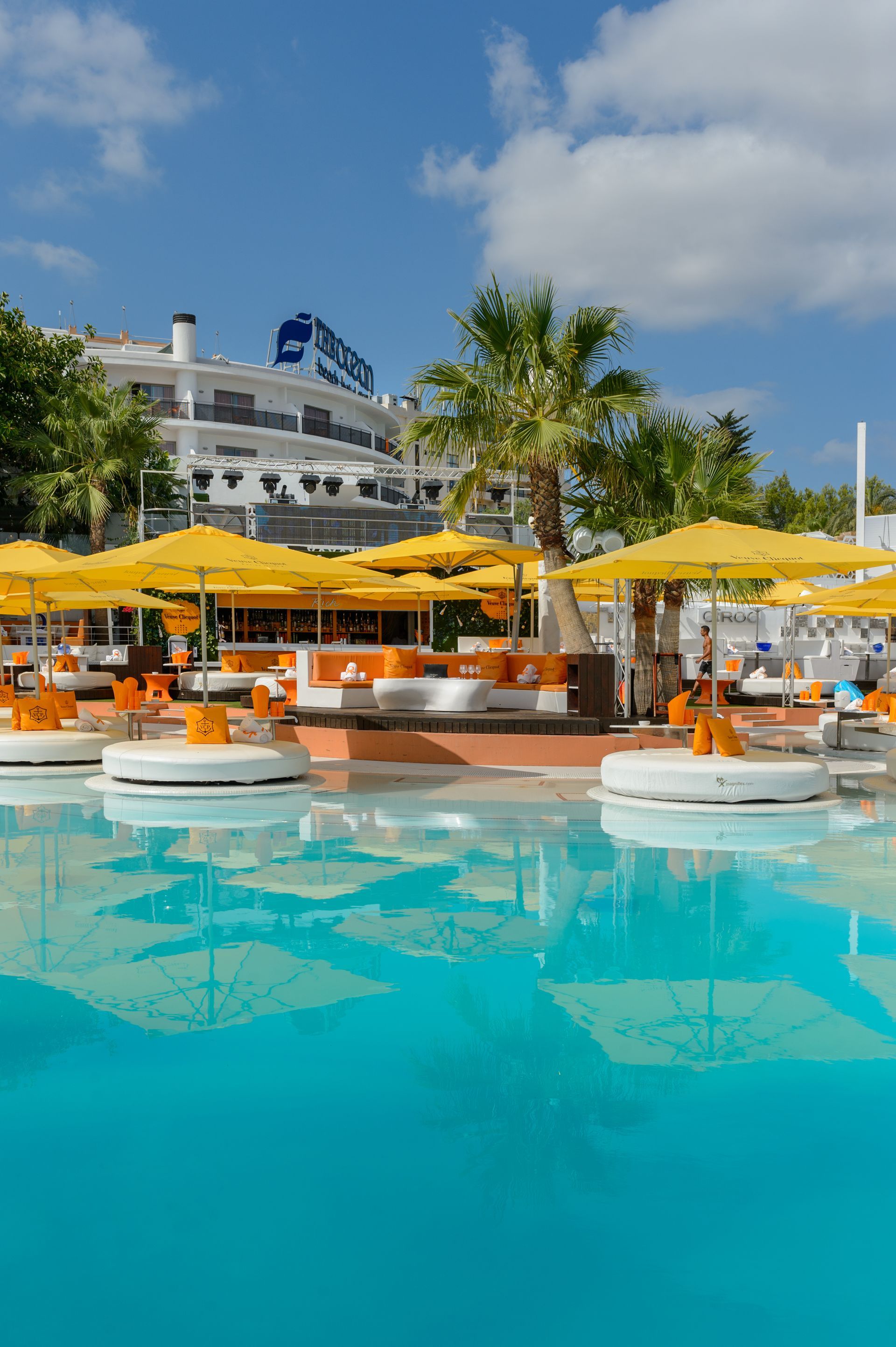 Complejo turístico junto a la piscina con agua turquesa, sombrillas amarillas y asientos blancos bajo un cielo azul.