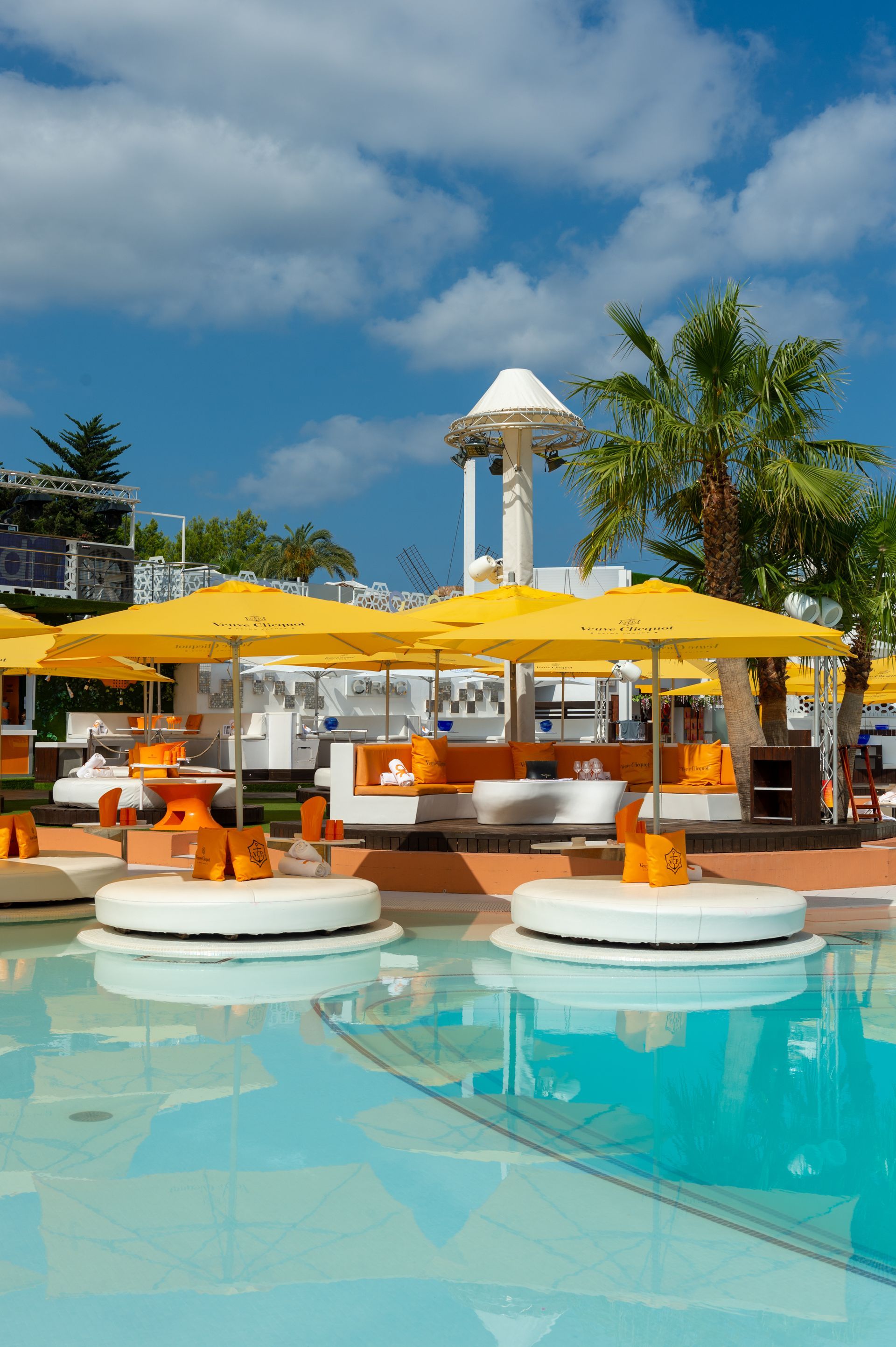 Zona de salón junto a la piscina con sombrillas amarillas, asientos blancos y una torre bajo un cielo azul.