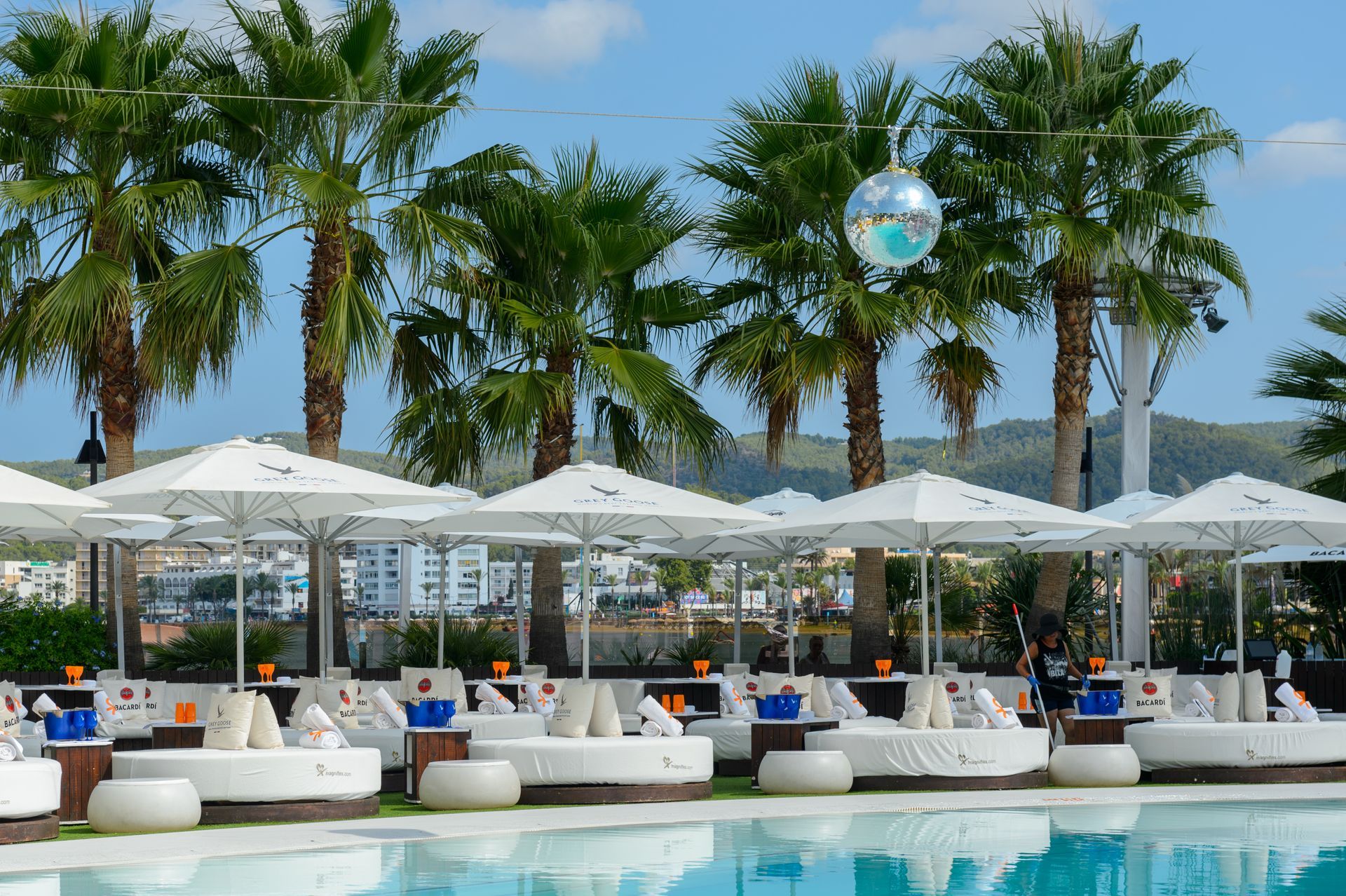 Escena junto a la piscina con tumbonas blancas, sombrillas, palmeras y una piscina azul bajo un cielo soleado.