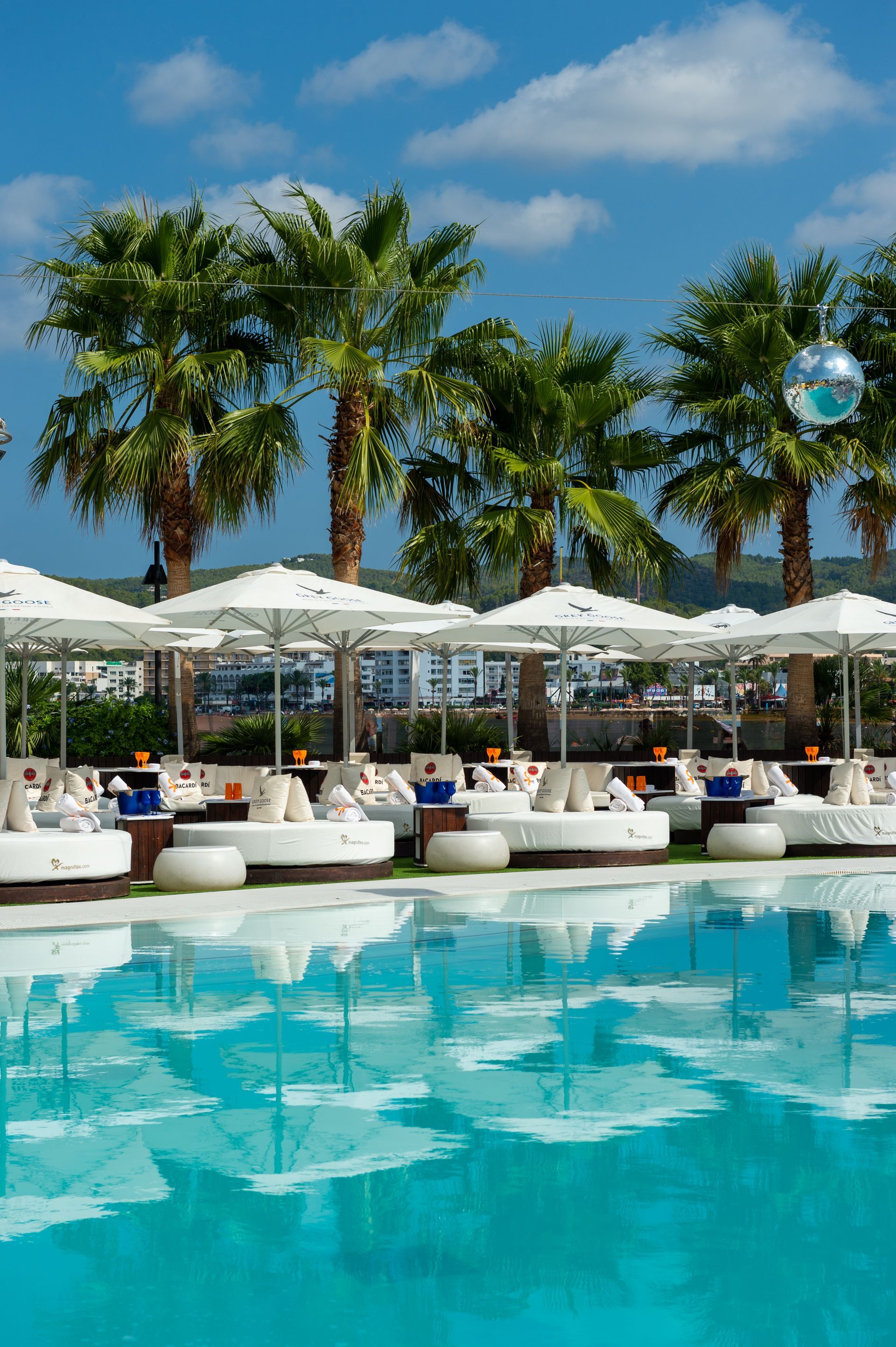Piscina con tumbonas, sombrillas blancas y palmeras bajo un cielo azul.