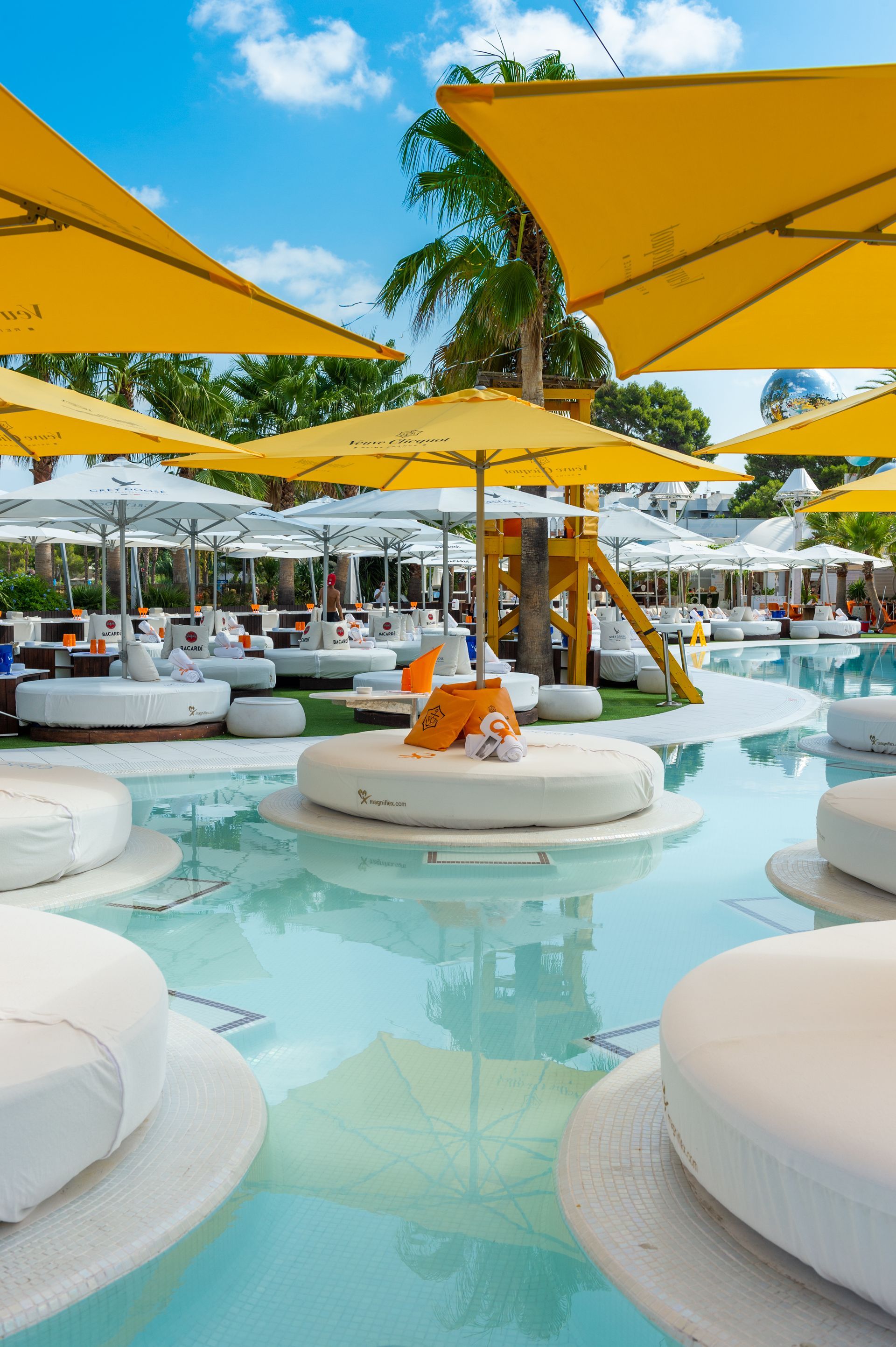 Zona de salón junto a la piscina con plataformas blancas, sombrillas amarillas y agua turquesa cristalina.
