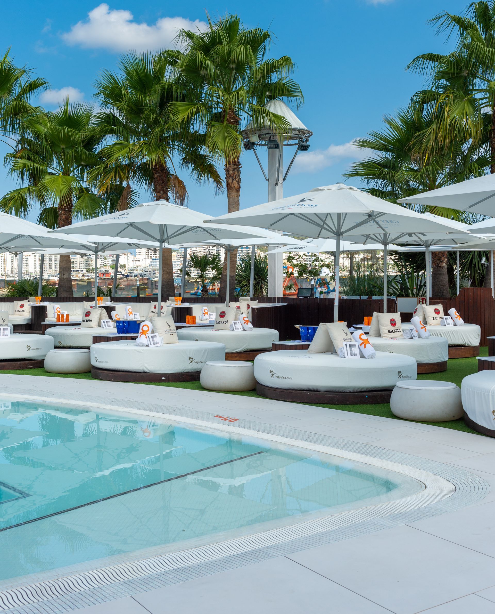 Zona de salón junto a la piscina con tumbonas blancas, sombrillas y palmeras.