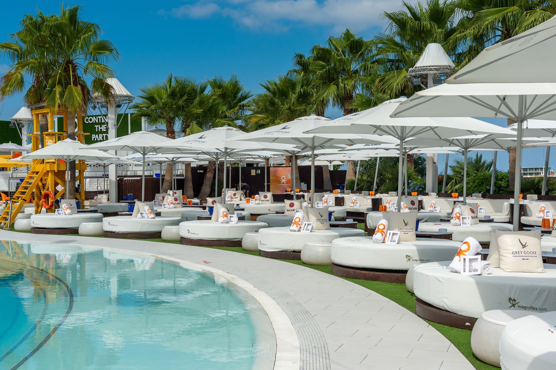 Salón junto a la piscina con tumbonas blancas, sombrillas y palmeras bajo un cielo azul.