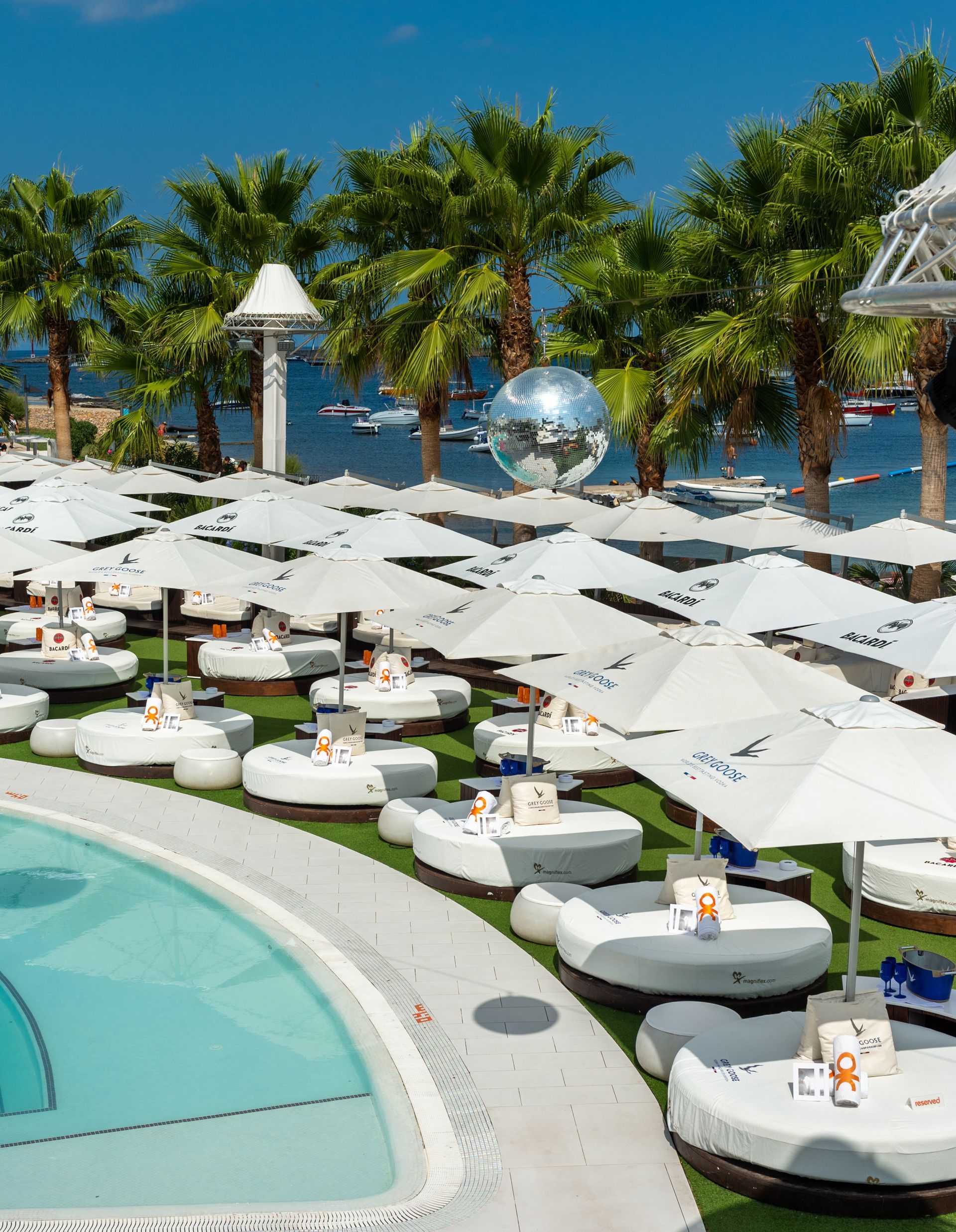 Zona de salón junto a la piscina con tumbonas blancas, sombrillas y una bola de discoteca, junto a una piscina y palmeras.
