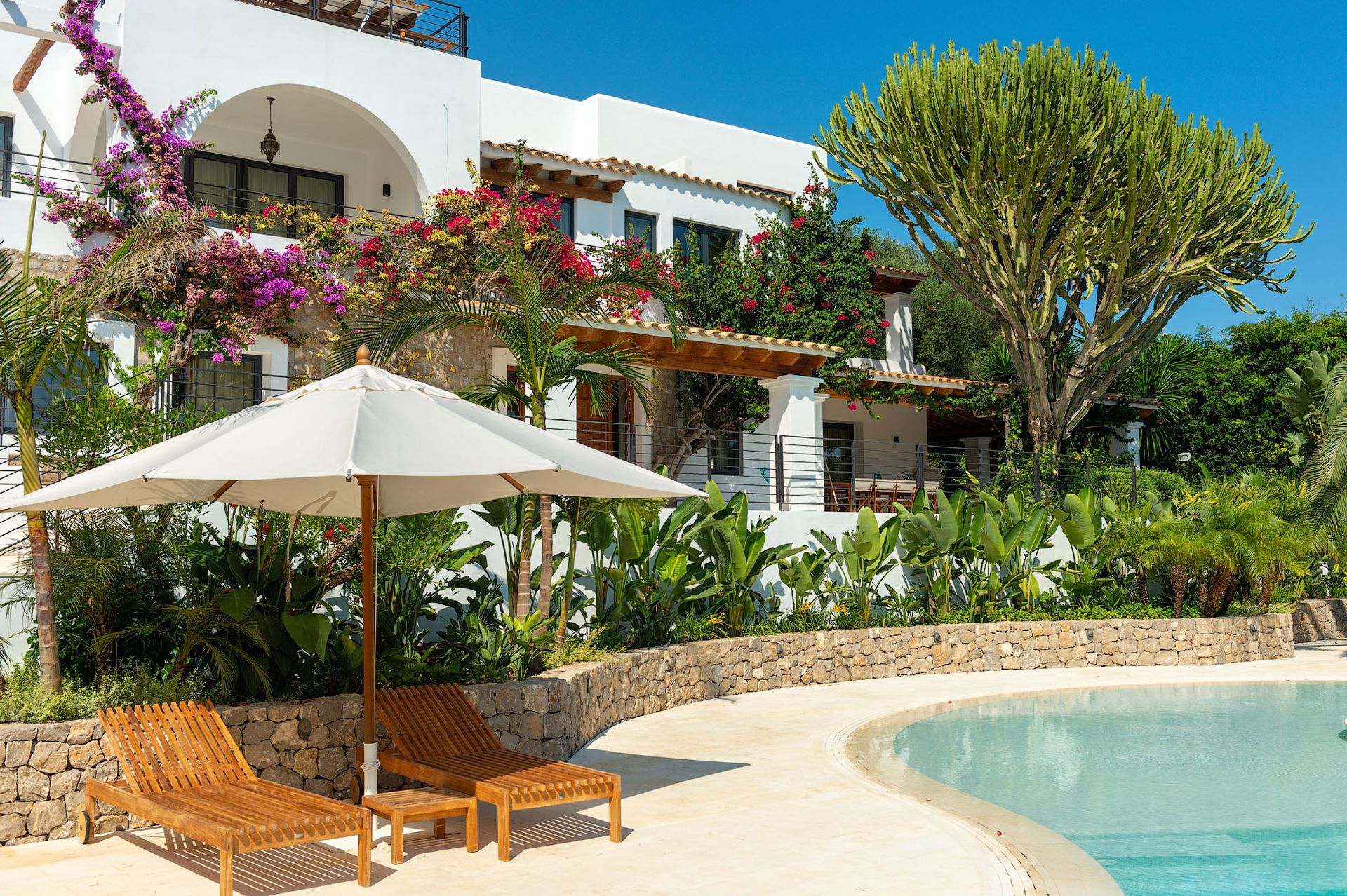 Dos tumbonas de madera bajo una sombrilla junto a la piscina. Villa blanca con plantas en flor al fondo.