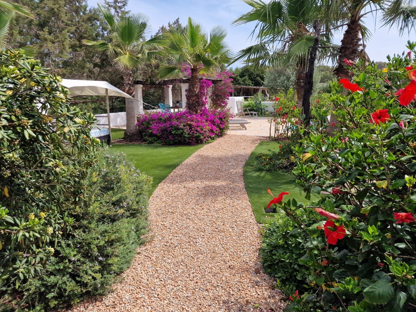 El camino de grava se curva a través de un jardín con follaje verde, flores rosadas y una estructura blanca en el fondo.