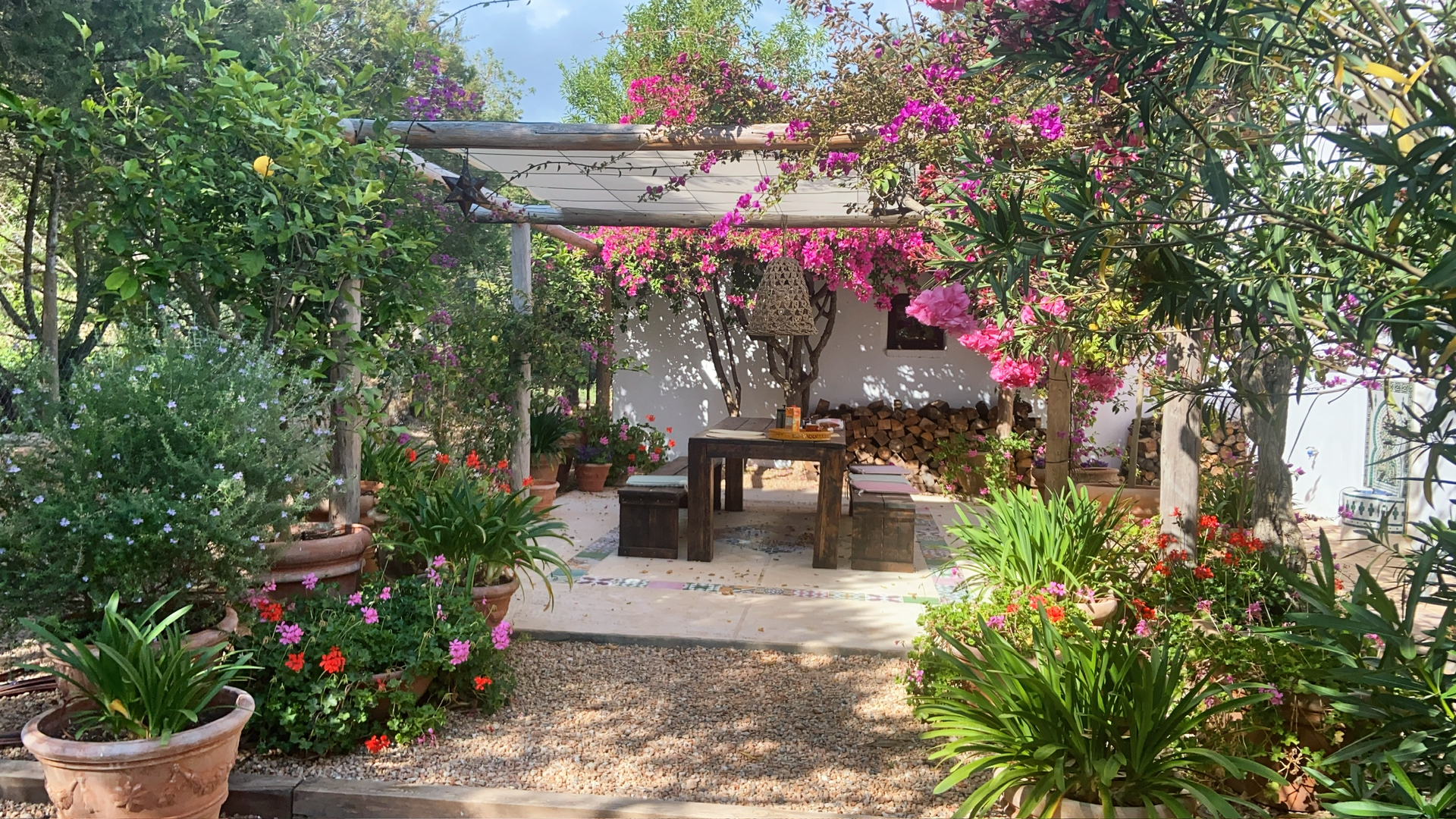 Área de comedor al aire libre rodeada de exuberante vegetación, enredaderas en flor y macetas de terracota.