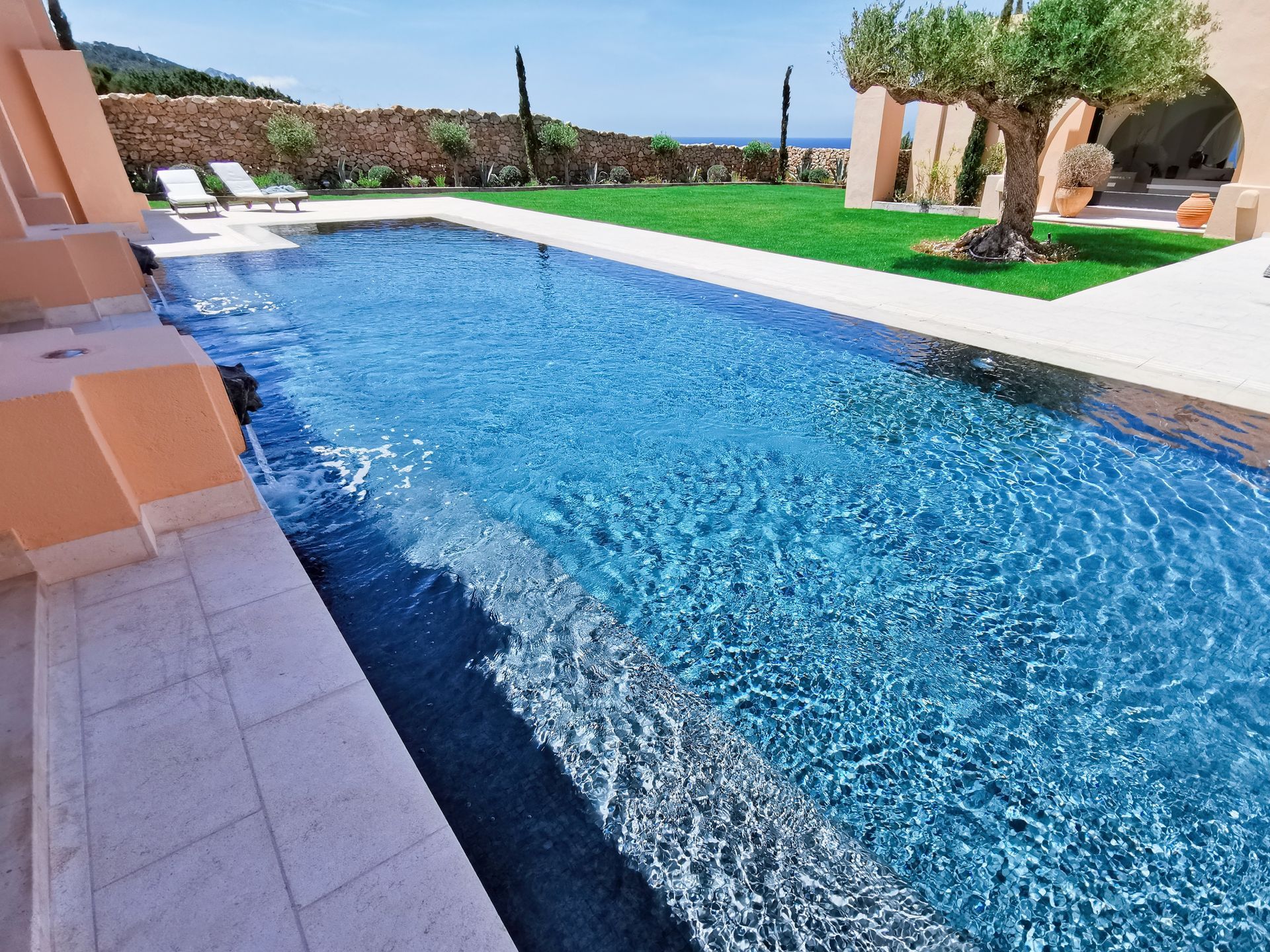 Piscina rectangular larga, agua azul, escalones beige, césped verde, cielo azul, casa al fondo.