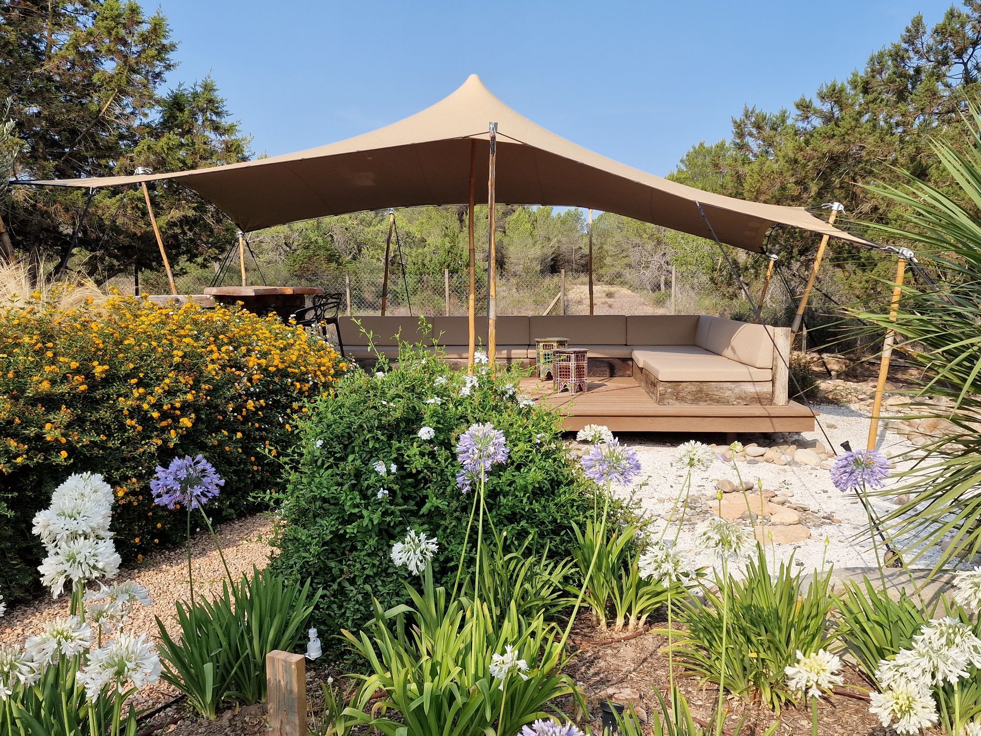 Tienda de campaña beige con zona de estar en un entorno de jardín, rodeada de flores y árboles.