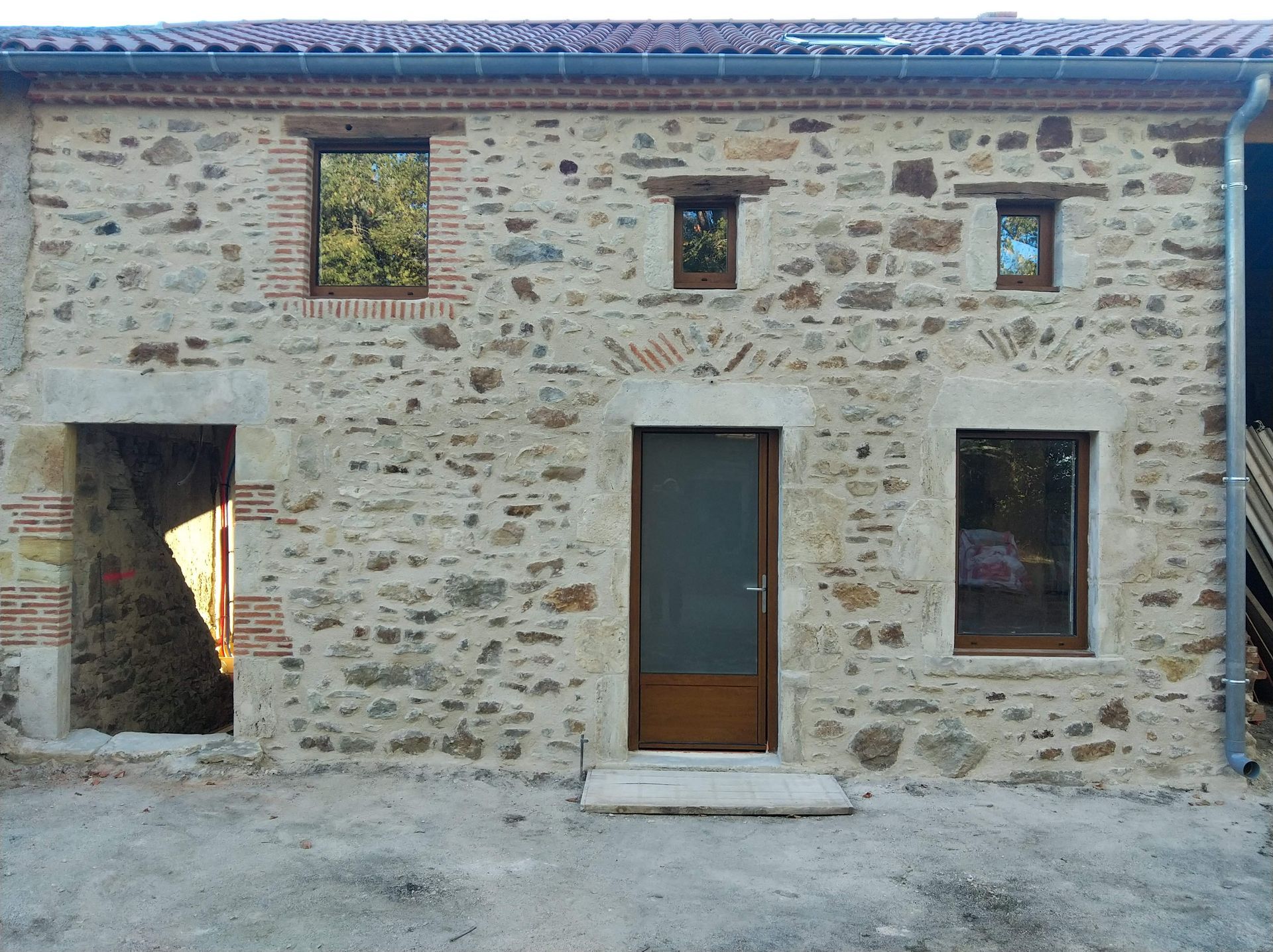 Façade de maison en pierre avec trois fenêtres, une porte centrale en bois et une cour gravillonnée.