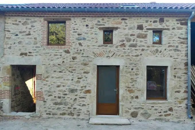 Façade de maison en pierre avec trois fenêtres en partie haute, une porte centrale en bois et une grande ouverture sur la gauche.