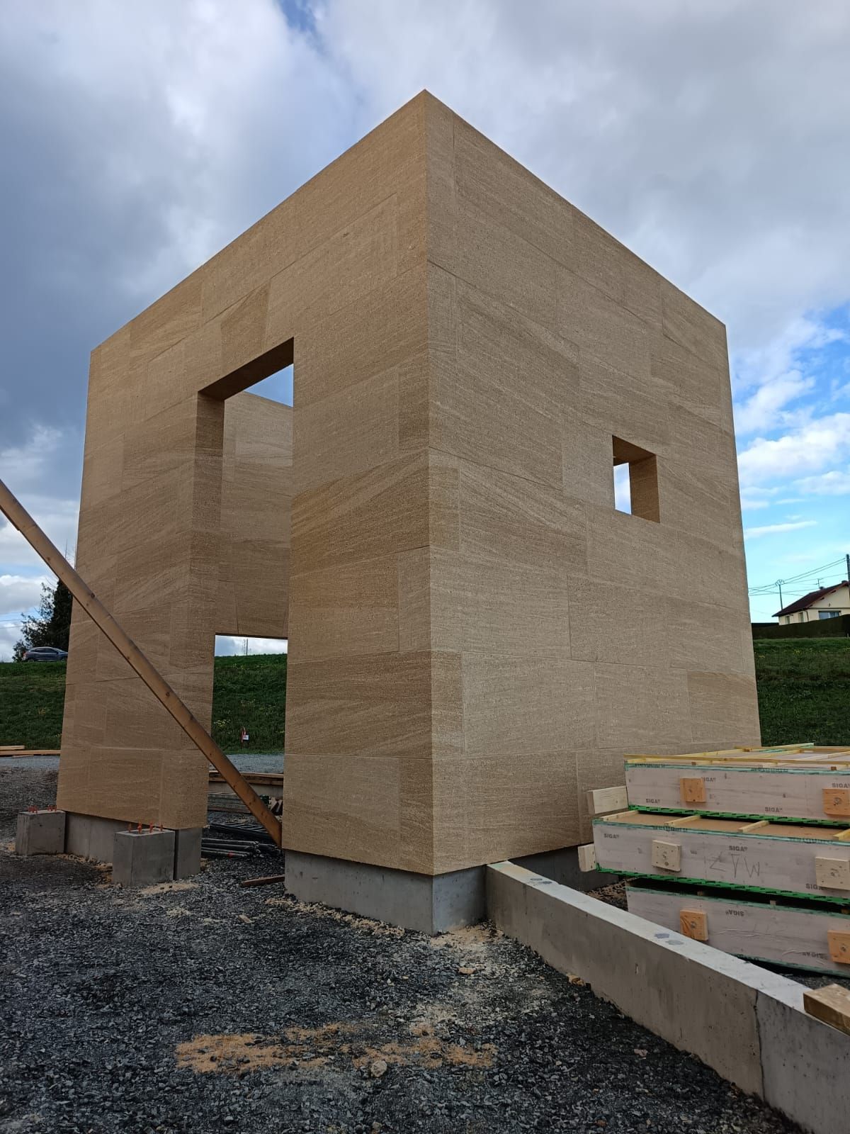 Ossature de maison en contreplaqué beige inachevée, en construction sur un terrain gravillonné, avec des ouvertures pour les fenêtres et des matériaux empilés à proximité.