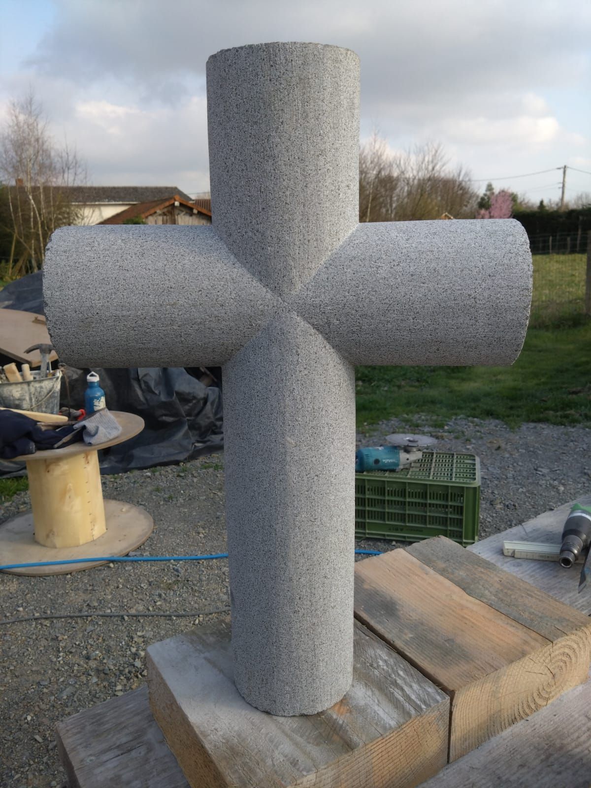 Sculpture en mousse grise en forme de croix, installée à l'extérieur sur un socle en béton dans la cour d'un atelier.