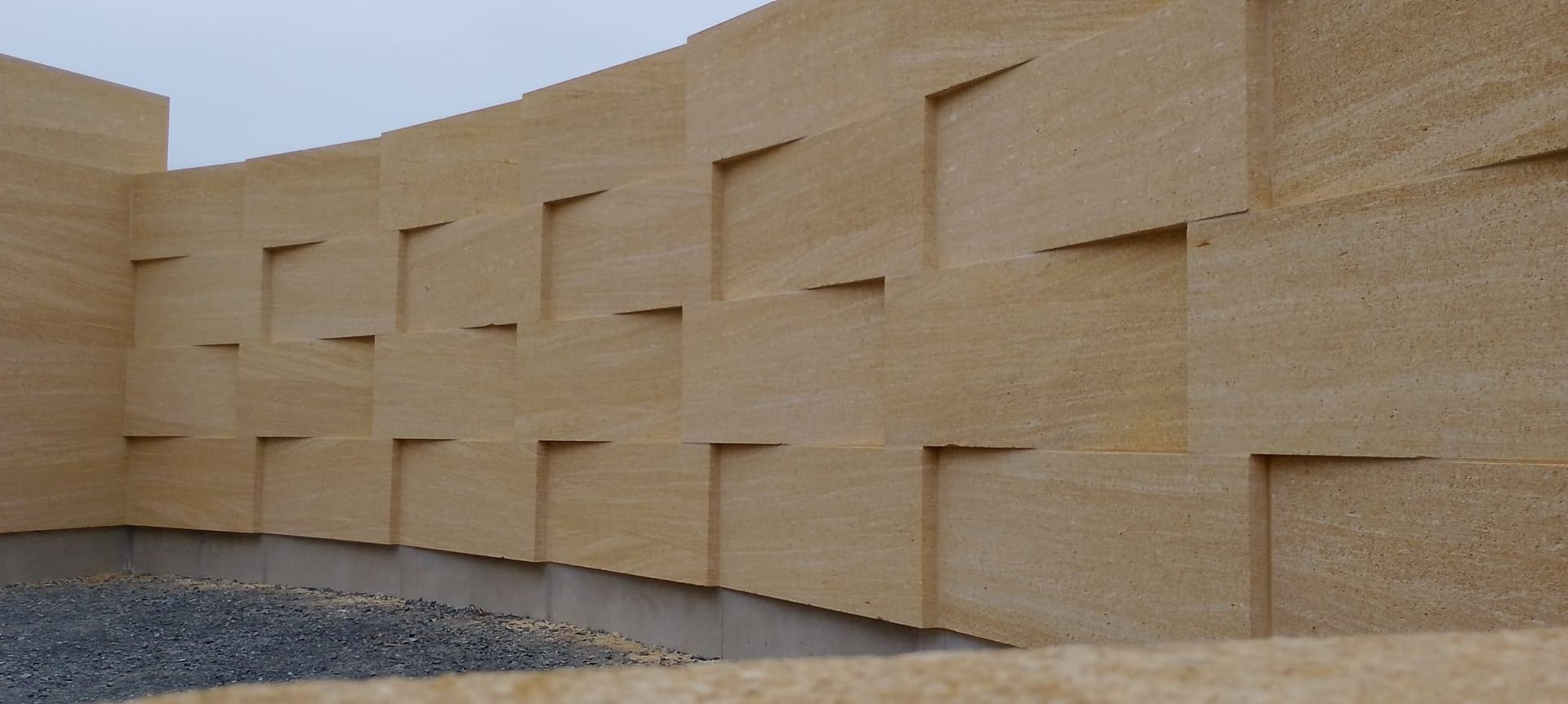 Mur incurvé en pierre beige avec des blocs étagés à côté d'un sol de gravier sous la lumière du jour