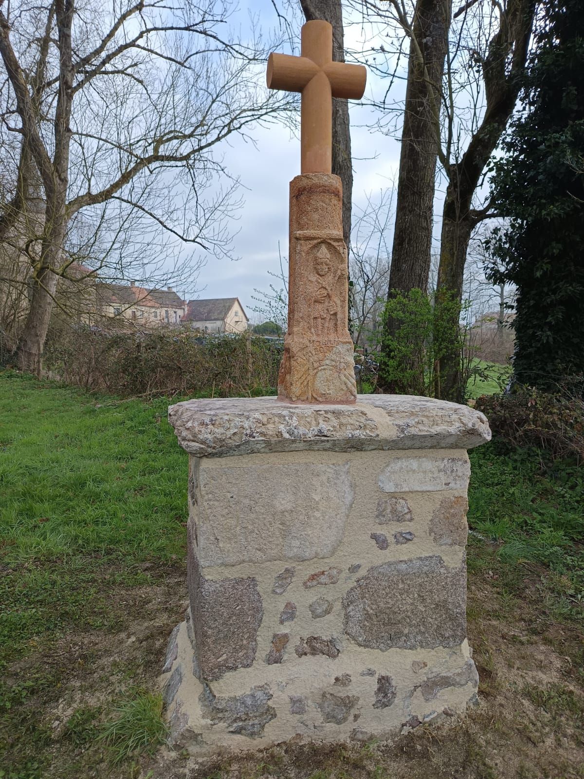 Croix de pierre sur un piédestal dans un espace vert extérieur avec des arbres et des maisons en arrière-plan
