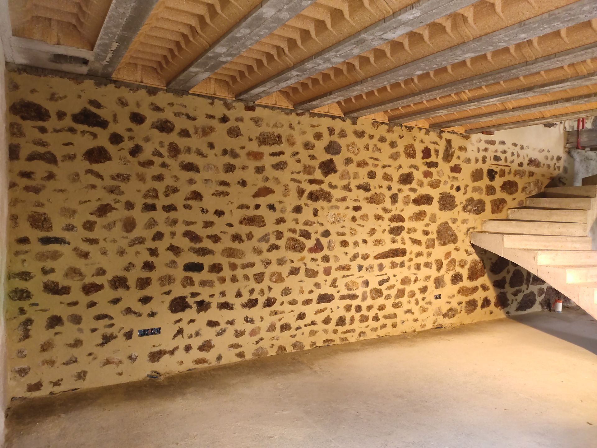 Sous-sol aux murs de pierre avec poutres apparentes au plafond et un escalier sur la droite