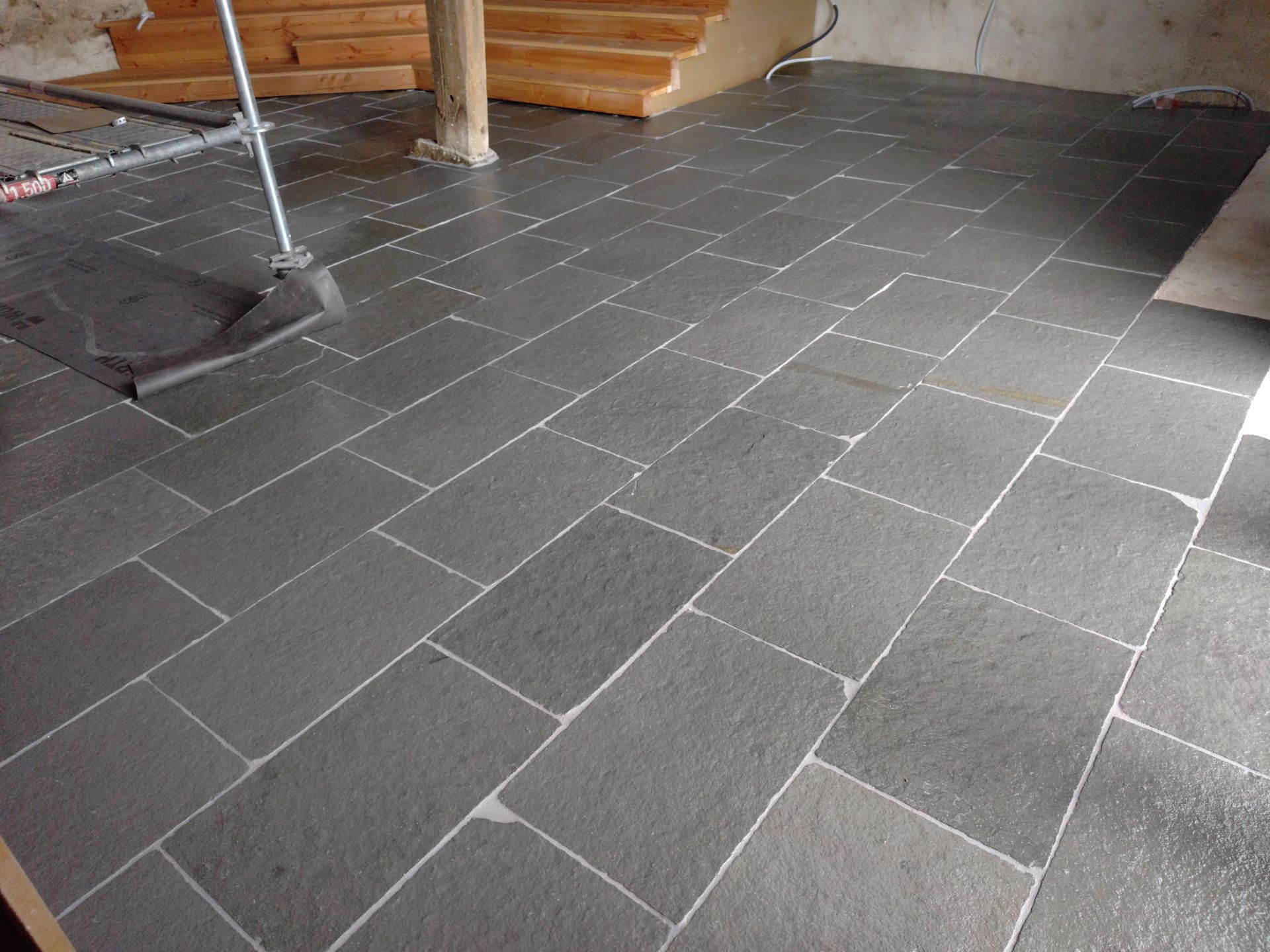 Sol carrelé en construction avec des carreaux gris, des joints blancs et un escalier en arrière-plan.