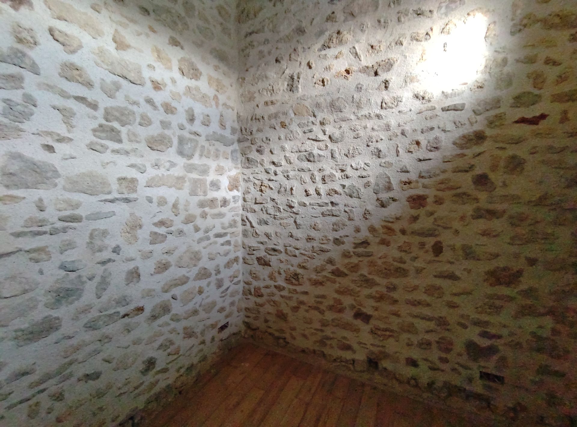 Pièce d'angle aux murs de pierre, avec un sol en bois et la lumière du soleil qui entre par le coin supérieur droit.