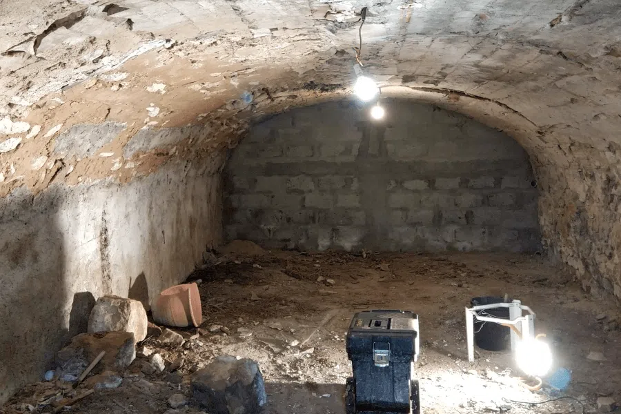 Cave en pierre brute avec plafond voûté, ampoule suspendue et outils et débris éparpillés.