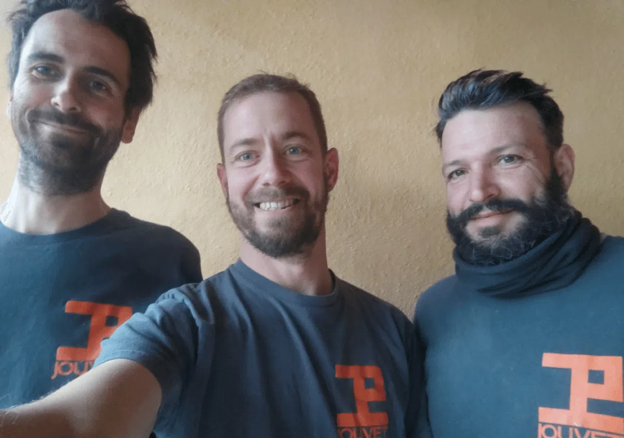 Trois hommes souriants en chemises sombres posent à l'intérieur, devant un mur beige.