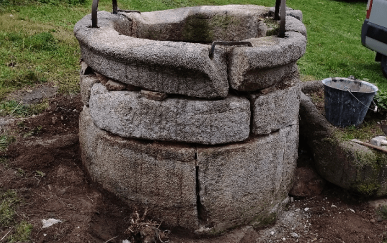 Un puits en pierres empilées se trouve dans une cour herbeuse, avec un seau à proximité.