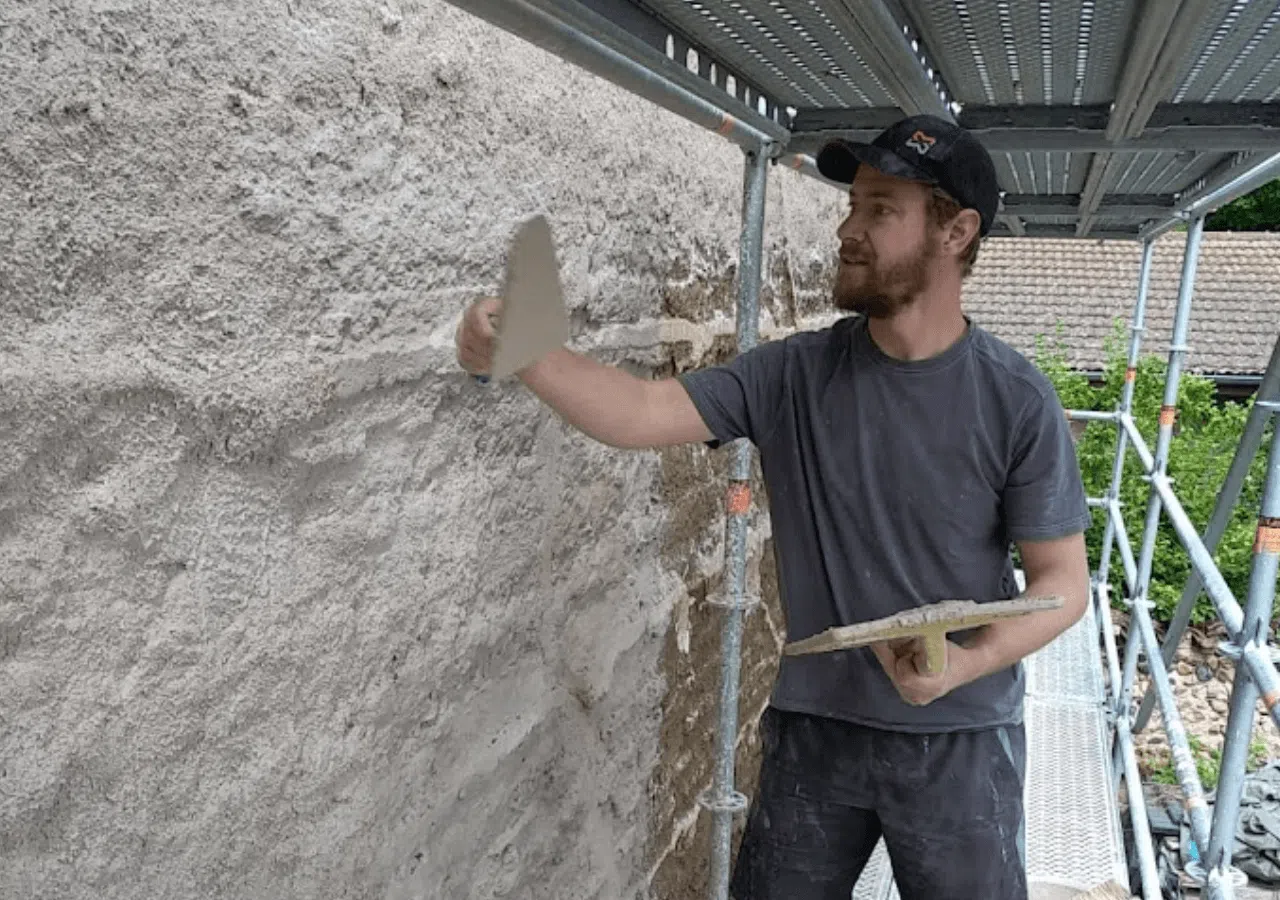 Un ouvrier enduit un mur extérieur brut sous un échafaudage métallique, tenant une truelle et une truelle.