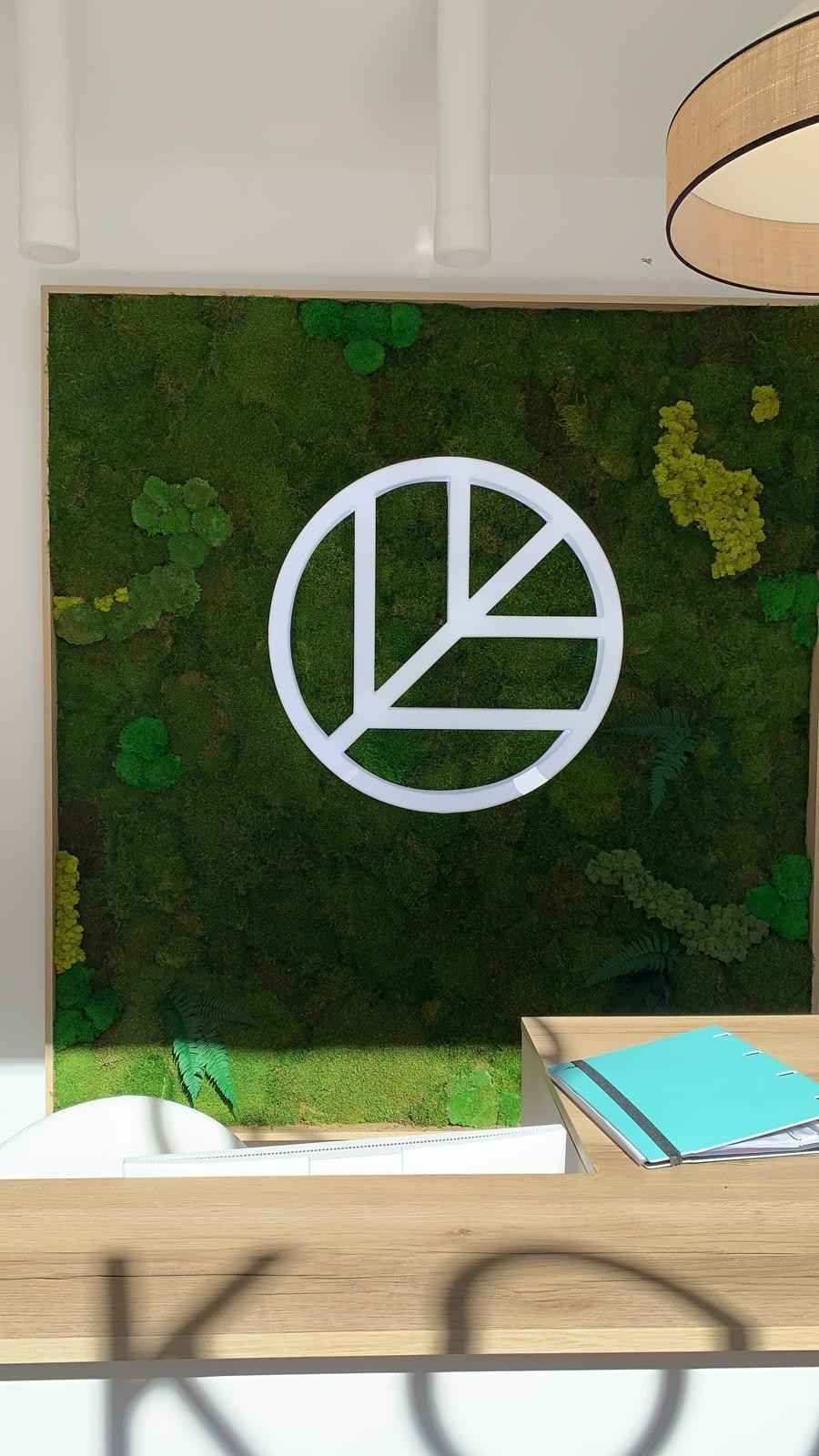 Pared de musgo verde con un logotipo circular blanco sobre un escritorio con papeles y una carpeta azul.