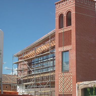 Edificio de ladrillo en construcción, andamios, ventanas de vidrio, torre con aberturas arqueadas, cielo azul claro.