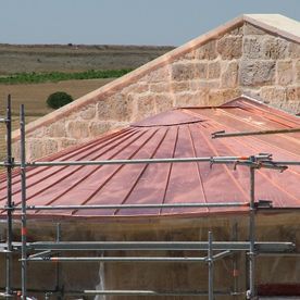 Edificio con techo de cobre en construcción; paredes de piedra beige, andamios y paisaje rural al fondo.