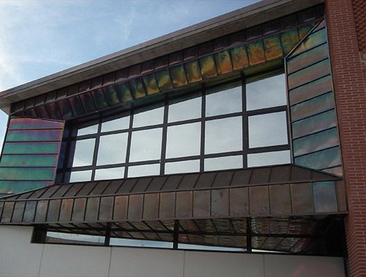 Exterior del edificio con gran ventanal, molduras de cobre y pared de ladrillo. El cielo despejado se refleja en el cristal.
